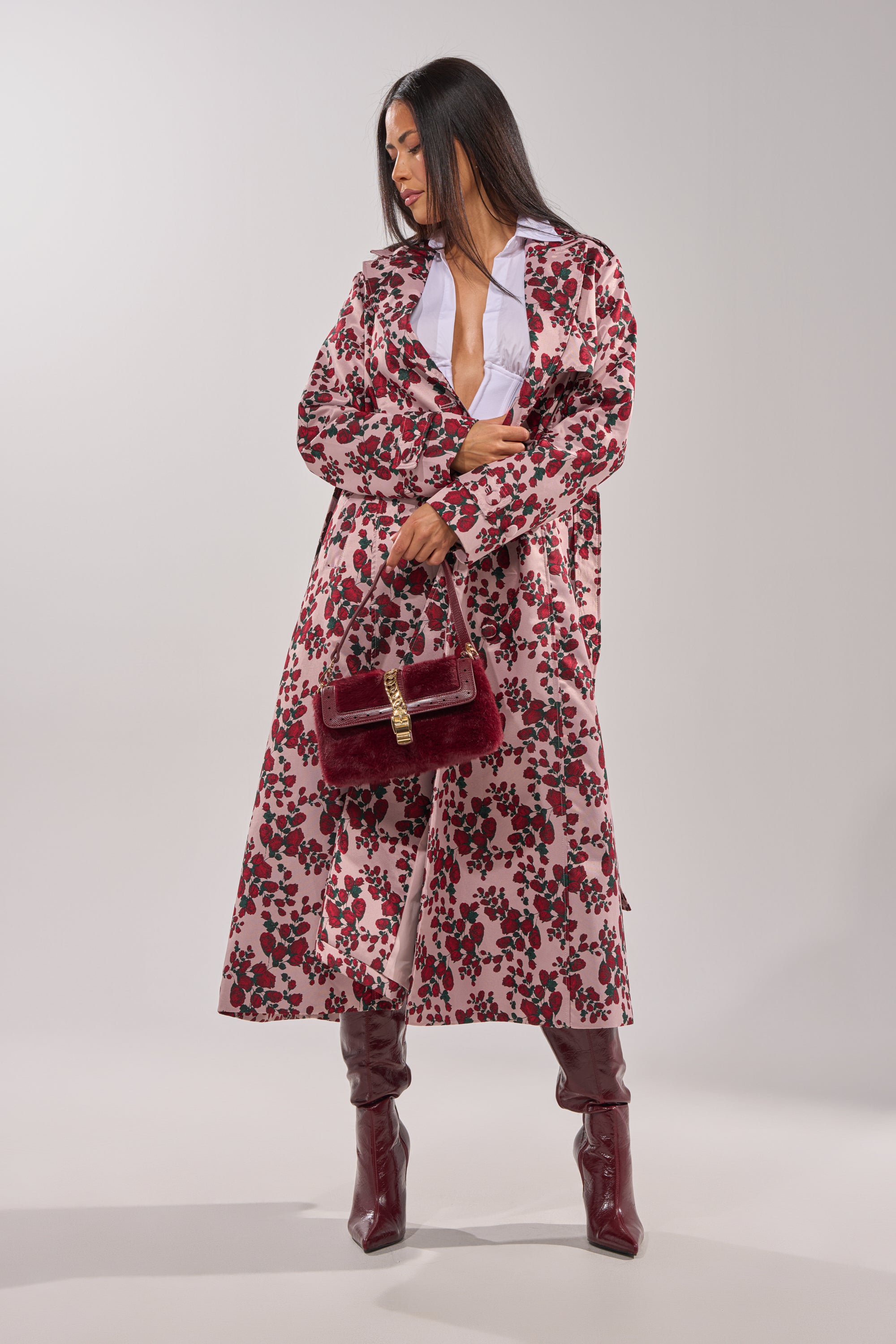 A woman poses in the BUY ME ROSES JACQUARD TRENCH, styled with deep red knee-high boots and a matching velvet handbag. She has long, straight dark hair and looks down while holding her coat open with one hand. 5