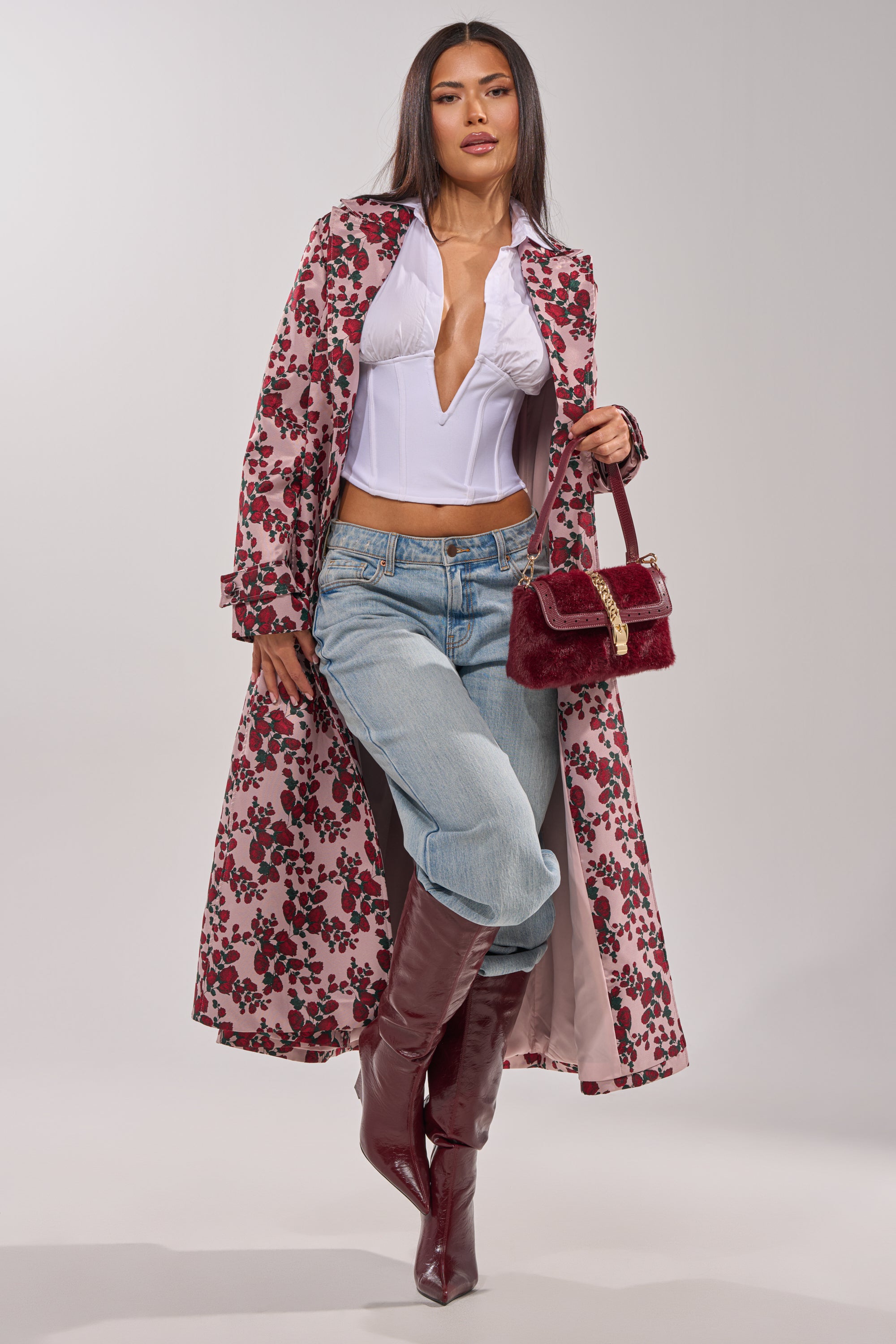 A woman poses in the BUY ME ROSES JACQUARD TRENCH with a white corset top, blue jeans, knee-high burgundy boots, and a matching burgundy handbag. Her straight, long hair contrasts with the plain light background. 2