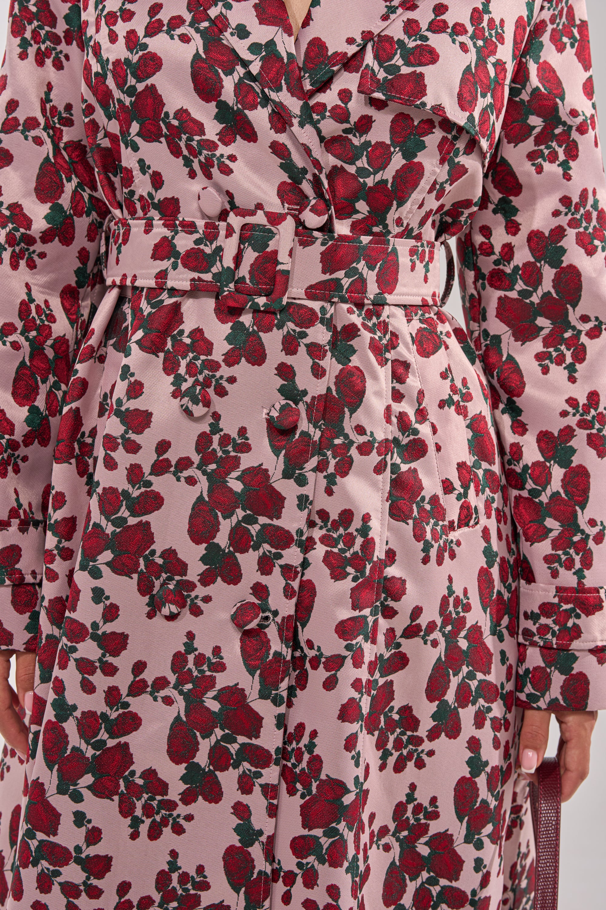 A close-up of the BUY ME ROSES JACQUARD TRENCH, a pink belted trench coat with a red and green floral pattern, highlighting its detailed fabric and belt—ideal as chic women's transitional outerwear. 11