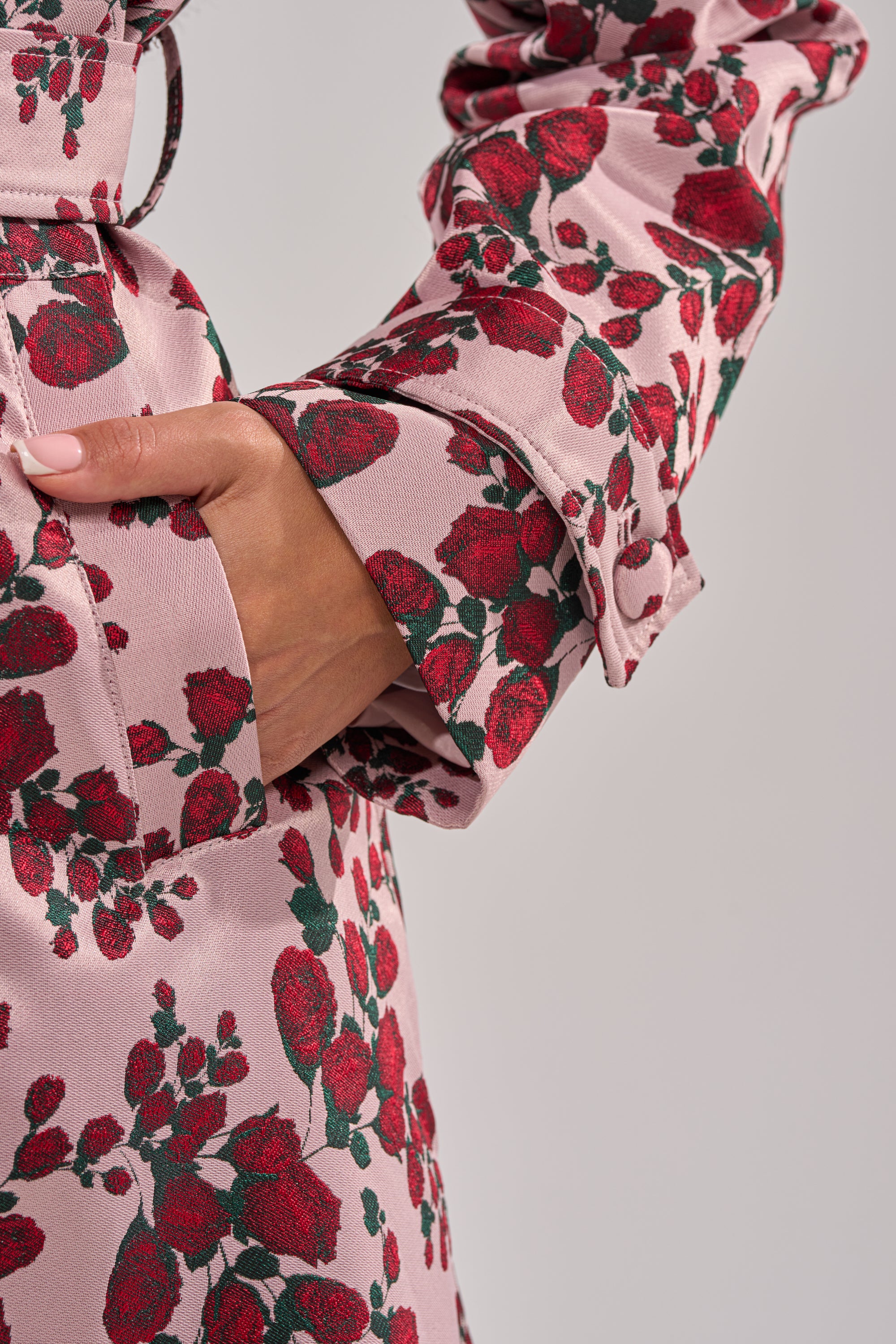 A close-up of a hand in the pocket of the BUY ME ROSES JACQUARD TRENCH shows its pink belted design with red rose print, soft fabric texture, and elegant sleeve and pocket details—perfect women’s transitional outerwear. 12