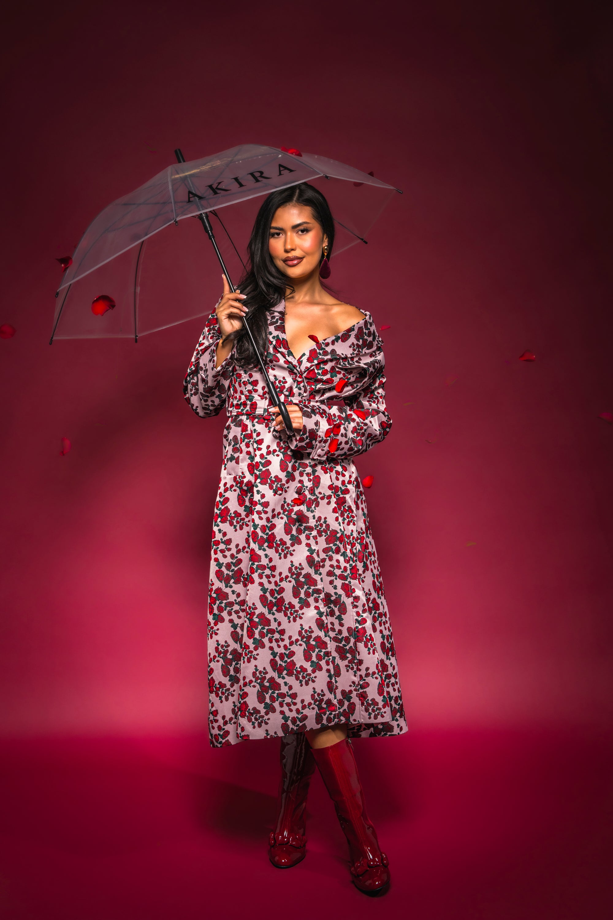 Wearing the BUY ME ROSES JACQUARD TRENCH, a woman stands confidently with a clear umbrella against a red background with petals, pairing the chic floral print coat with red boots and showcasing stylish transitional outerwear. 0