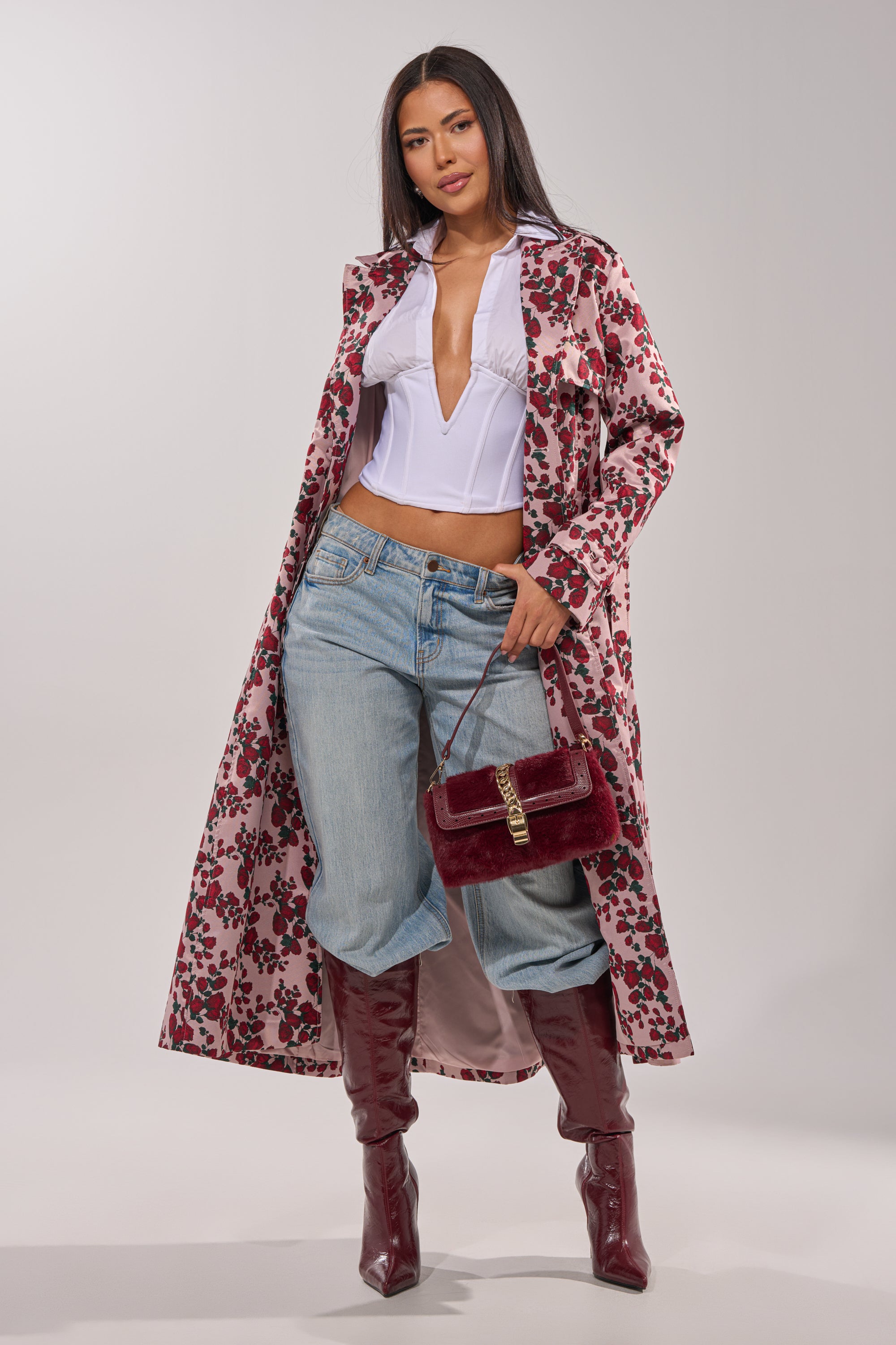 A woman poses in the BUY ME ROSES JACQUARD TRENCH, styled with a white low-cut blouse, light blue jeans, burgundy knee-high boots, and a matching handbag against a plain light background. 4