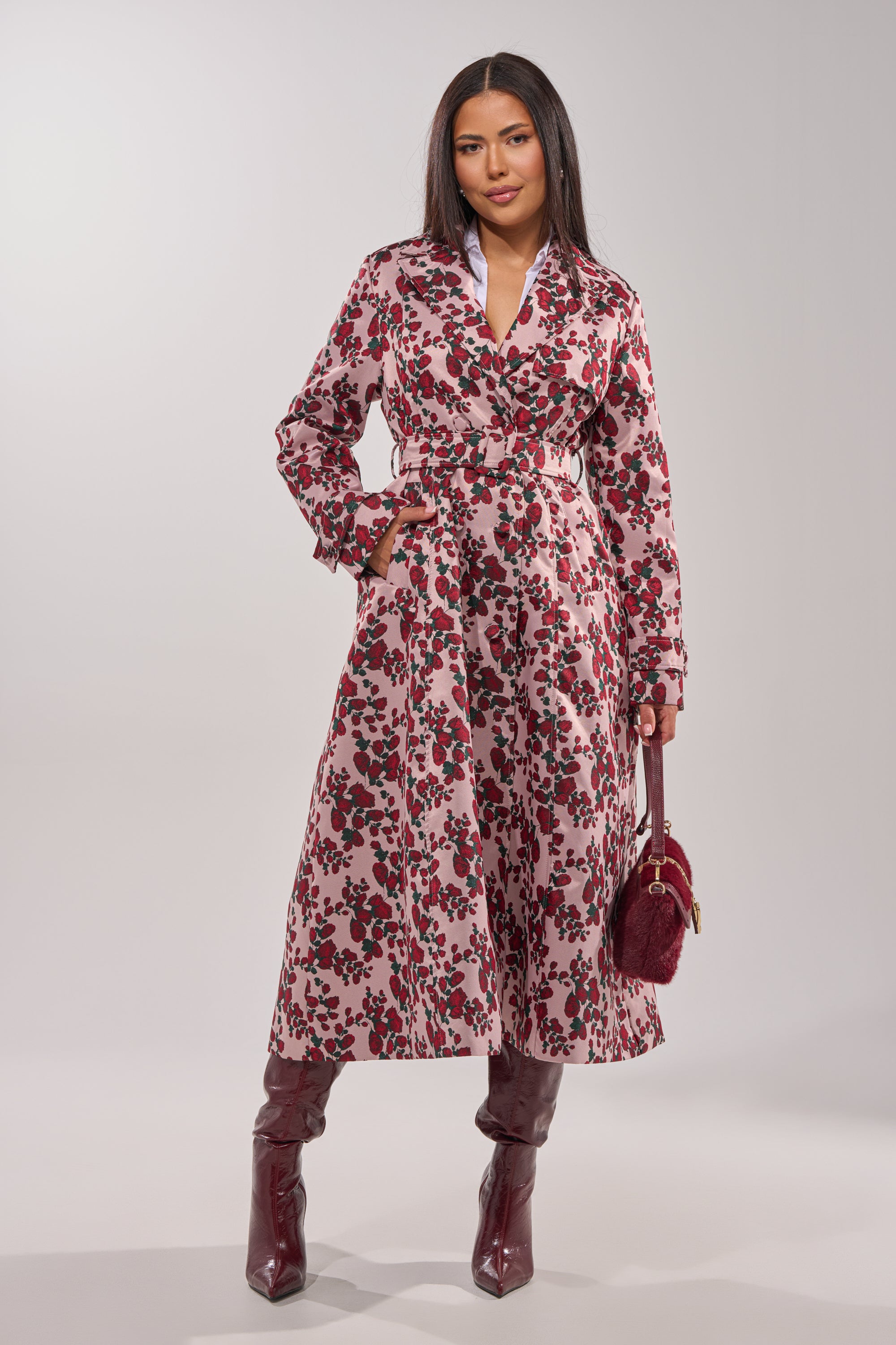 A woman with long dark hair poses against a plain light background, wearing the BUY ME ROSES JACQUARD TRENCH—a pink belted coat with a rose motif—paired with burgundy knee-high boots and a matching handbag. 6