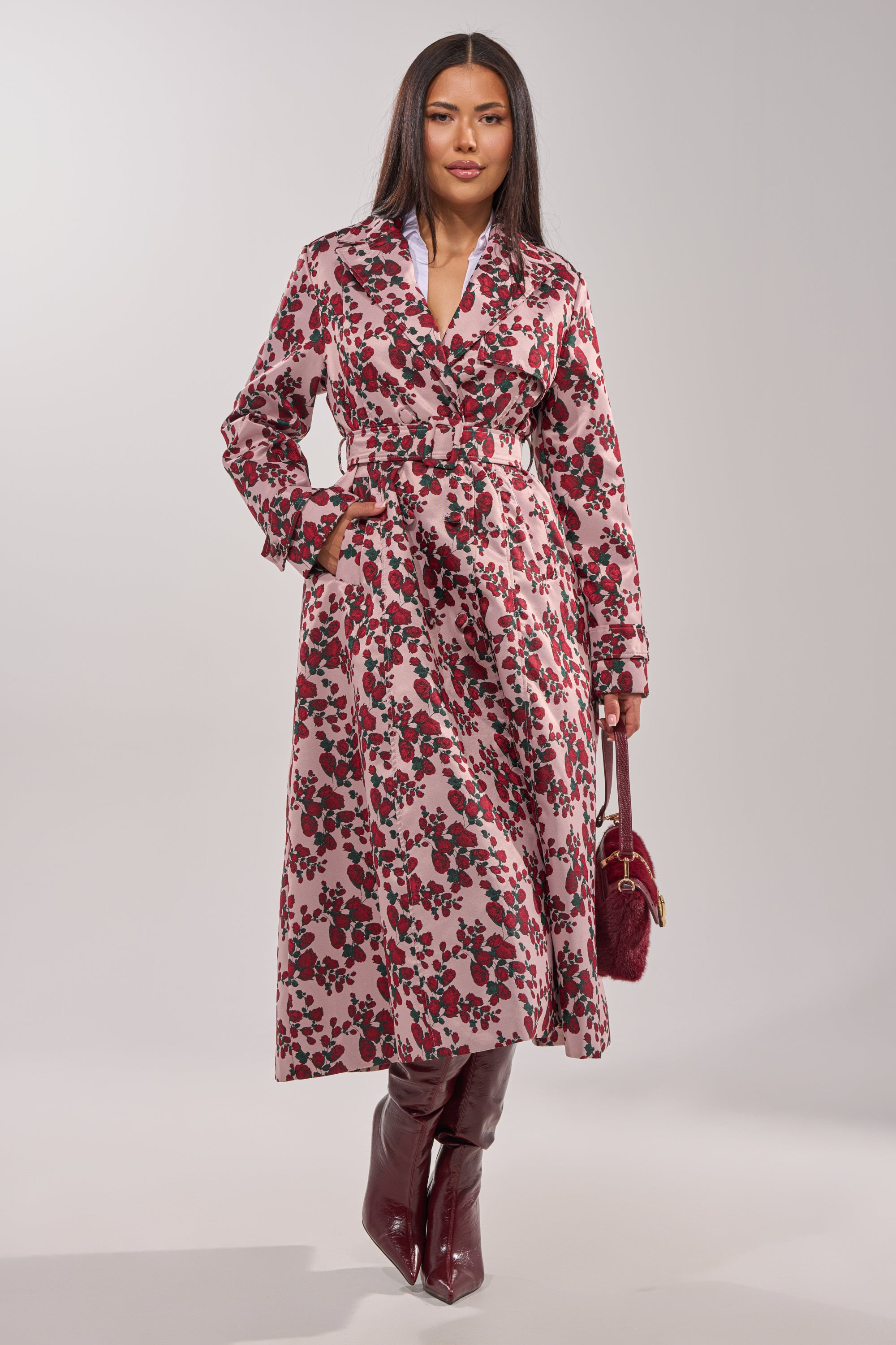 A woman exudes confidence wearing the BUY ME ROSES JACQUARD TRENCH with a pink belted floral design, styled with burgundy knee-high boots and a matching handbag, set against a light, neutral background. 7