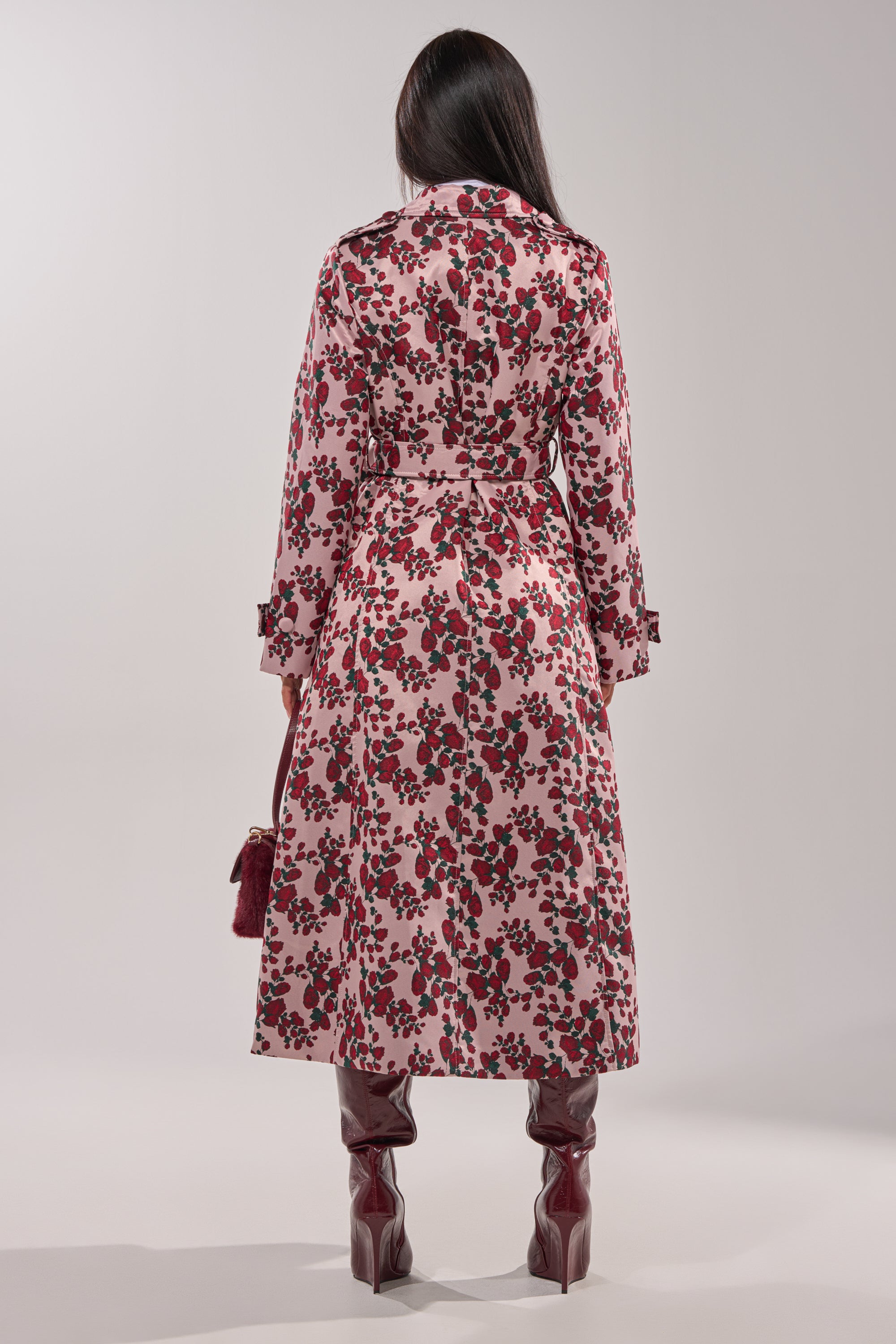 A person stands facing away in the BUY ME ROSES JACQUARD TRENCH, a pink belted coat with a rose motif, paired with glossy maroon knee-high boots and a matching small handbag against a plain light background. 9