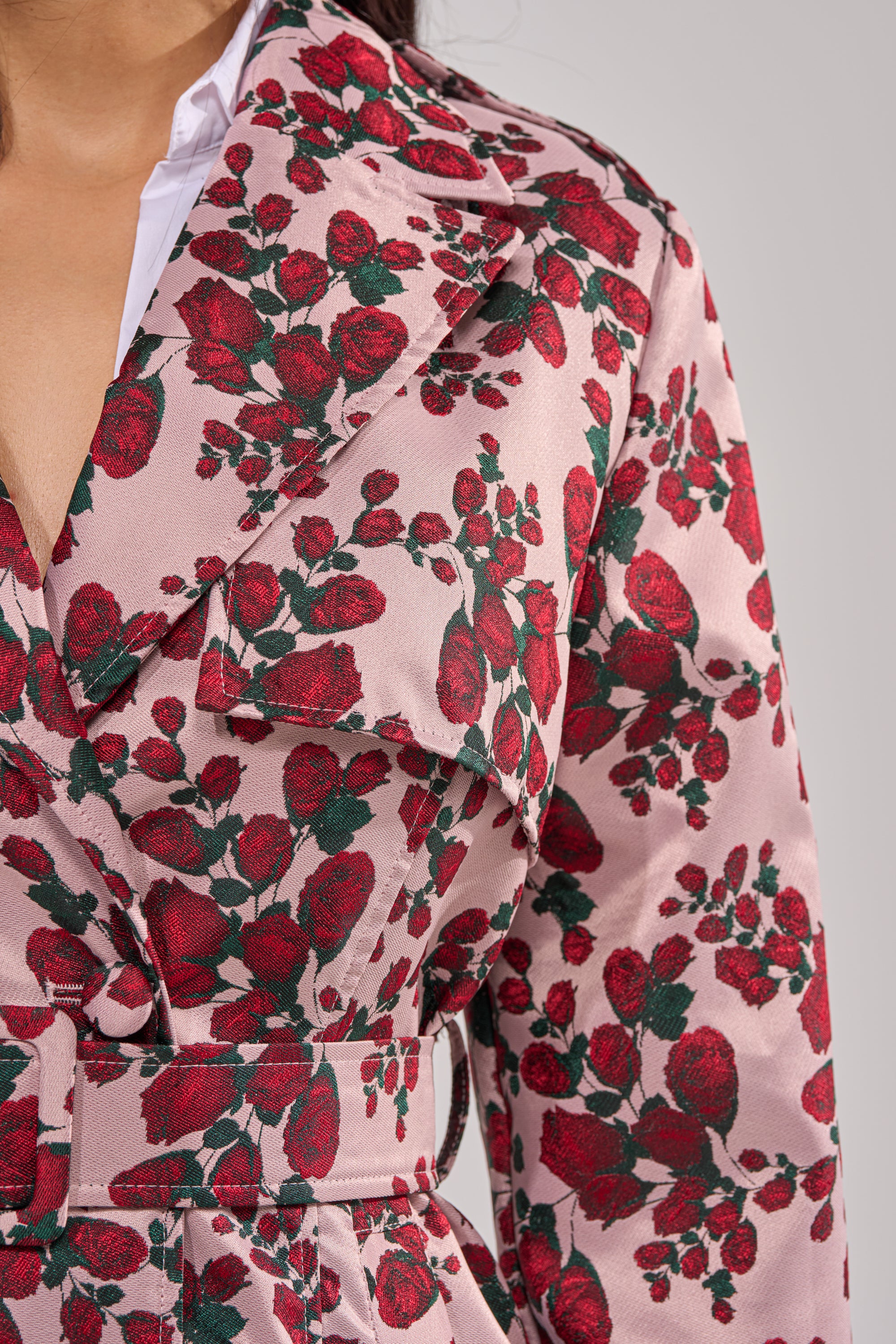 A close-up of a woman wearing the BUY ME ROSES JACQUARD TRENCH, a pink belted coat with a vibrant red and green rose motif, wide collar, matching belt, and the collar of a white shirt peeking underneath. 10