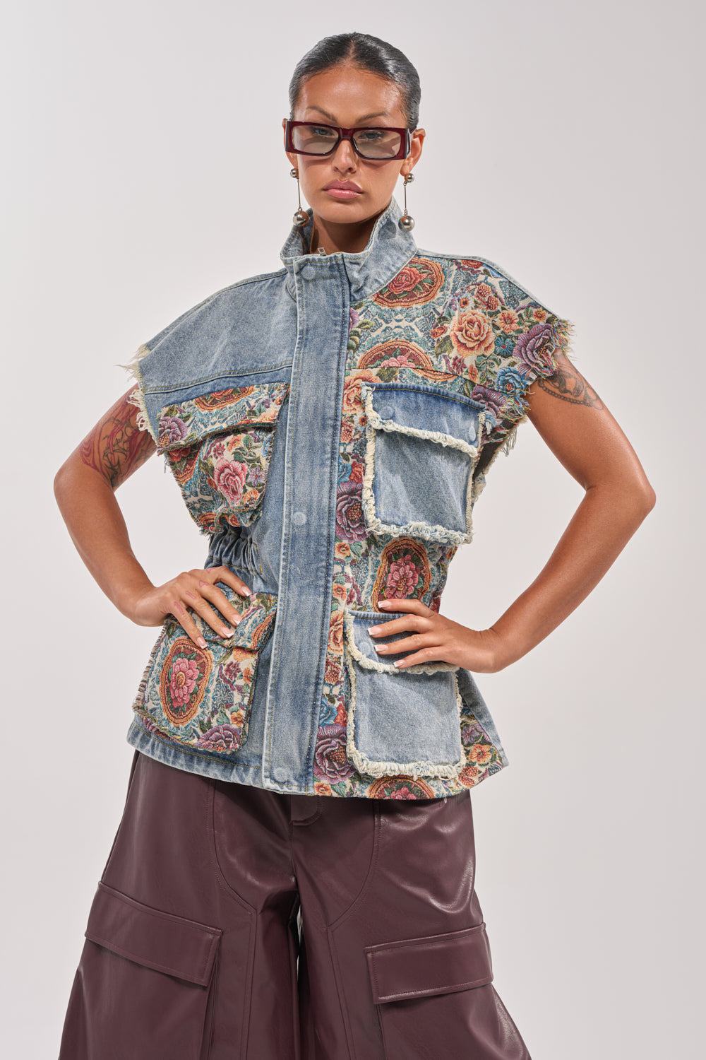 A woman poses confidently in the KAI PATCHWORK DENIM VEST, styled with bold glasses, statement earrings, and wide-leg brown pants for a trendy festival-ready look. 0