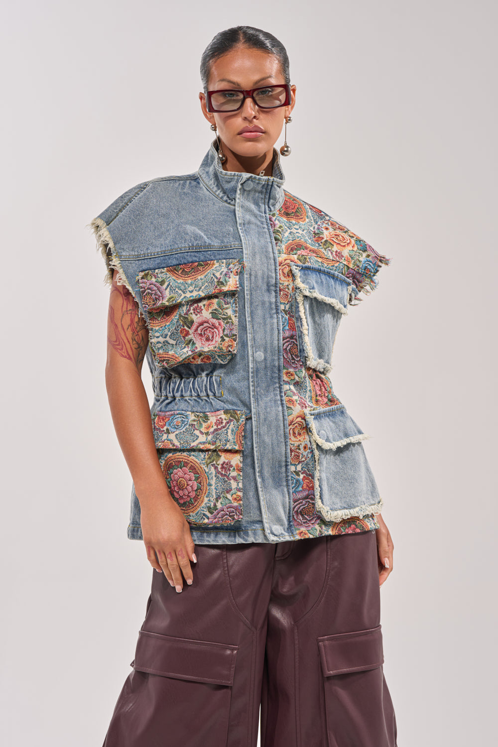 A woman exudes festival style in the KAI PATCHWORK DENIM VEST featuring floral tapestry patches, paired with brown wide-leg pants. With slicked-back hair, glasses, and statement earrings, she poses confidently with one hand in her pocket. 1