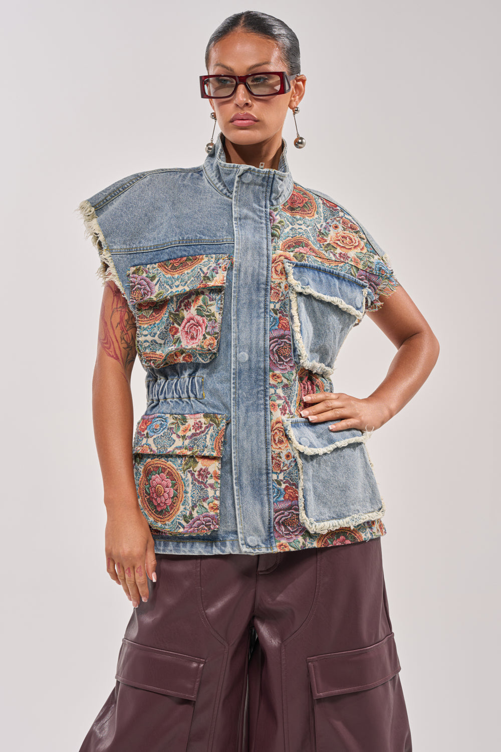 A person wearing dark sunglasses and bold earrings models the KAI PATCHWORK DENIM VEST with floral tapestry panels and large pockets, paired with wide-legged brown leather pants against a light background. 2