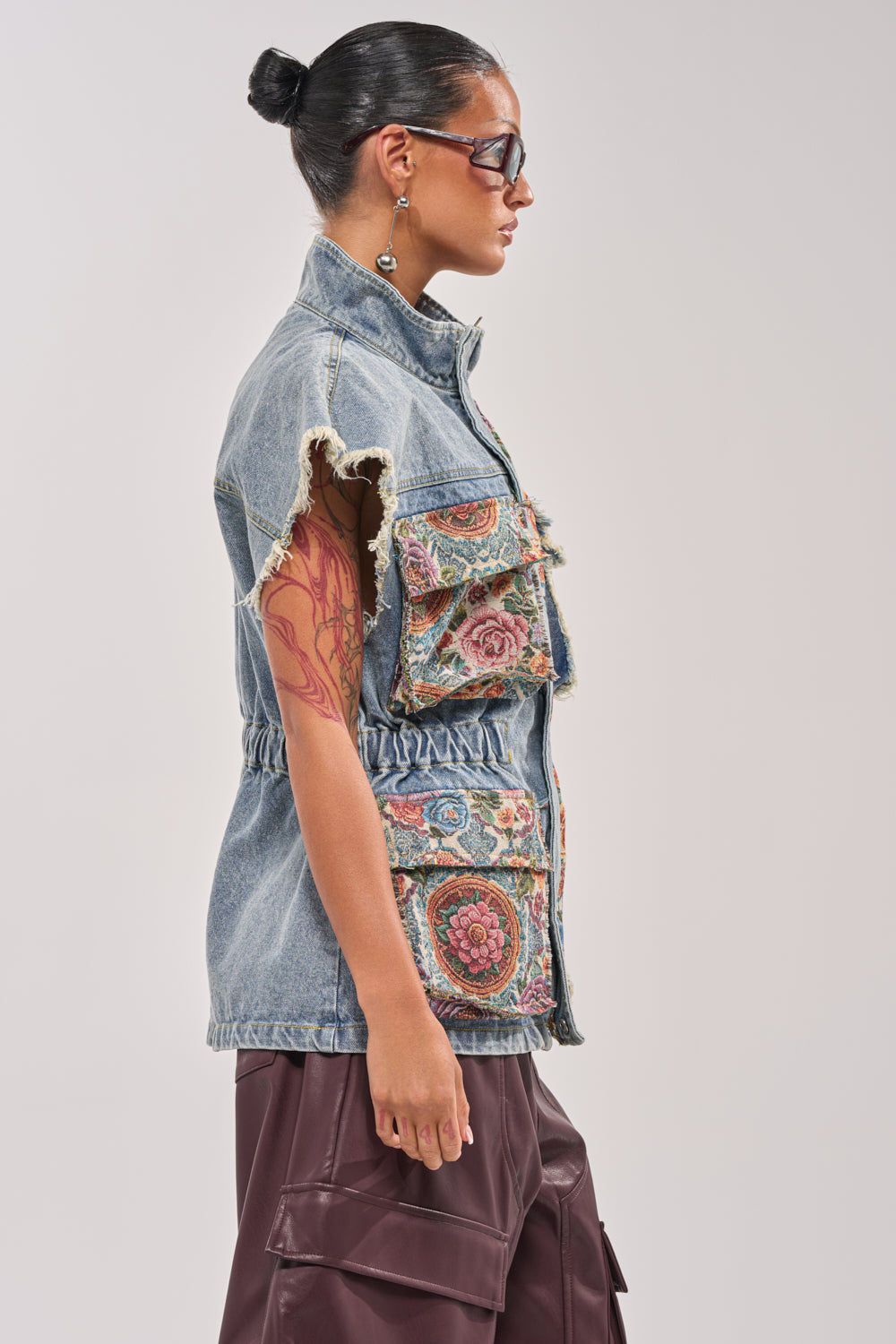 A person with dark hair in a bun, sunglasses, an earring, dark cargo pants, and the KAI PATCHWORK DENIM VEST featuring frayed edges and colorful patches stands in profile against a plain background. 3