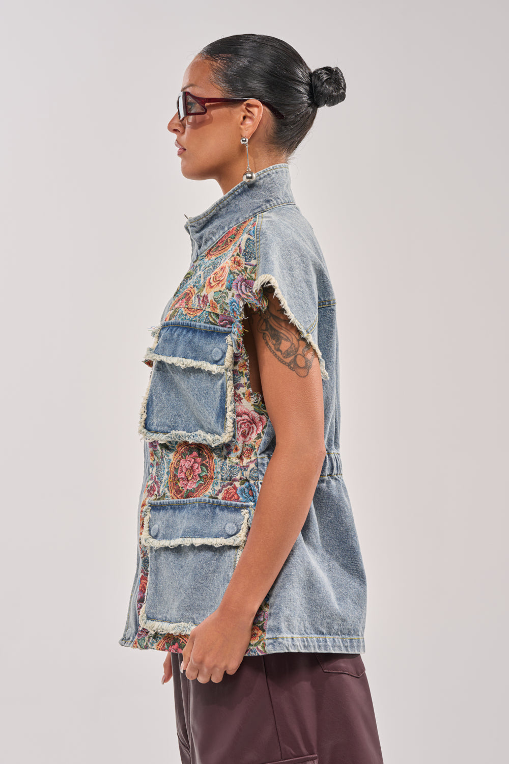 A person with dark hair in a bun, wearing sunglasses, the KAI PATCHWORK DENIM VEST with floral patterns and large pockets, and dark pants is shown in profile against a plain light background. 4