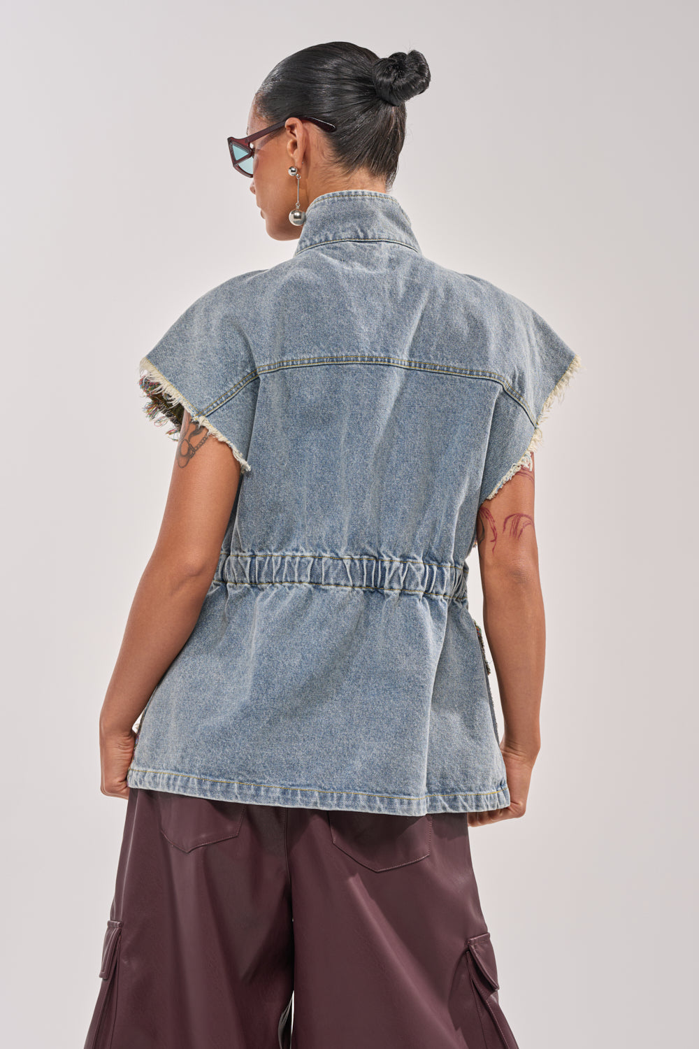 A person with hair in a neat bun, wearing glasses and the KAI PATCHWORK DENIM VEST with a cinched waist and dark loose pants, is shown from the back against a plain light background. 5