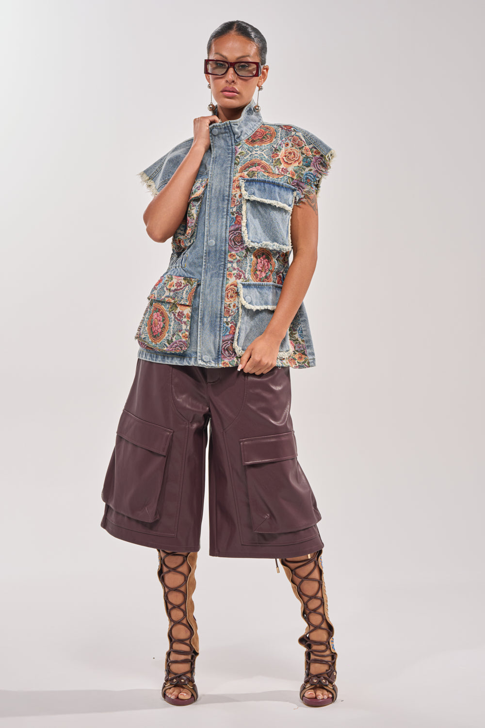 A woman in large sunglasses, the KAI PATCHWORK DENIM VEST with colorful embroidery and pockets, brown wide-leg cargo shorts, and knee-high lace-up sandals stands against a plain background. 6