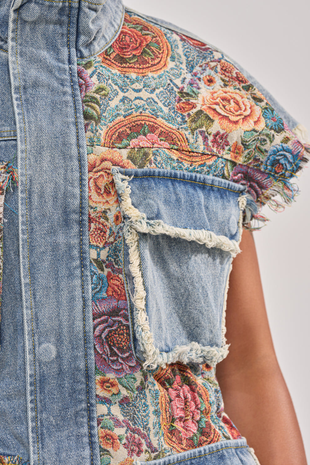 Close-up of the KAI PATCHWORK DENIM VEST: a sleeveless boho vest with a large frayed pocket and colorful floral tapestry pattern in pink, red, and blue—ideal for festival streetwear style. 7