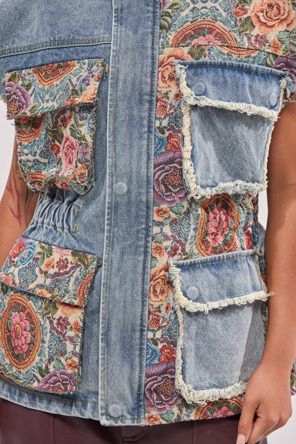 A close-up of the KAI PATCHWORK DENIM VEST, featuring floral tapestry panels and large frayed pockets. Styled with dark pants and a visible arm tattoo, it’s perfect for festival streetwear. 8