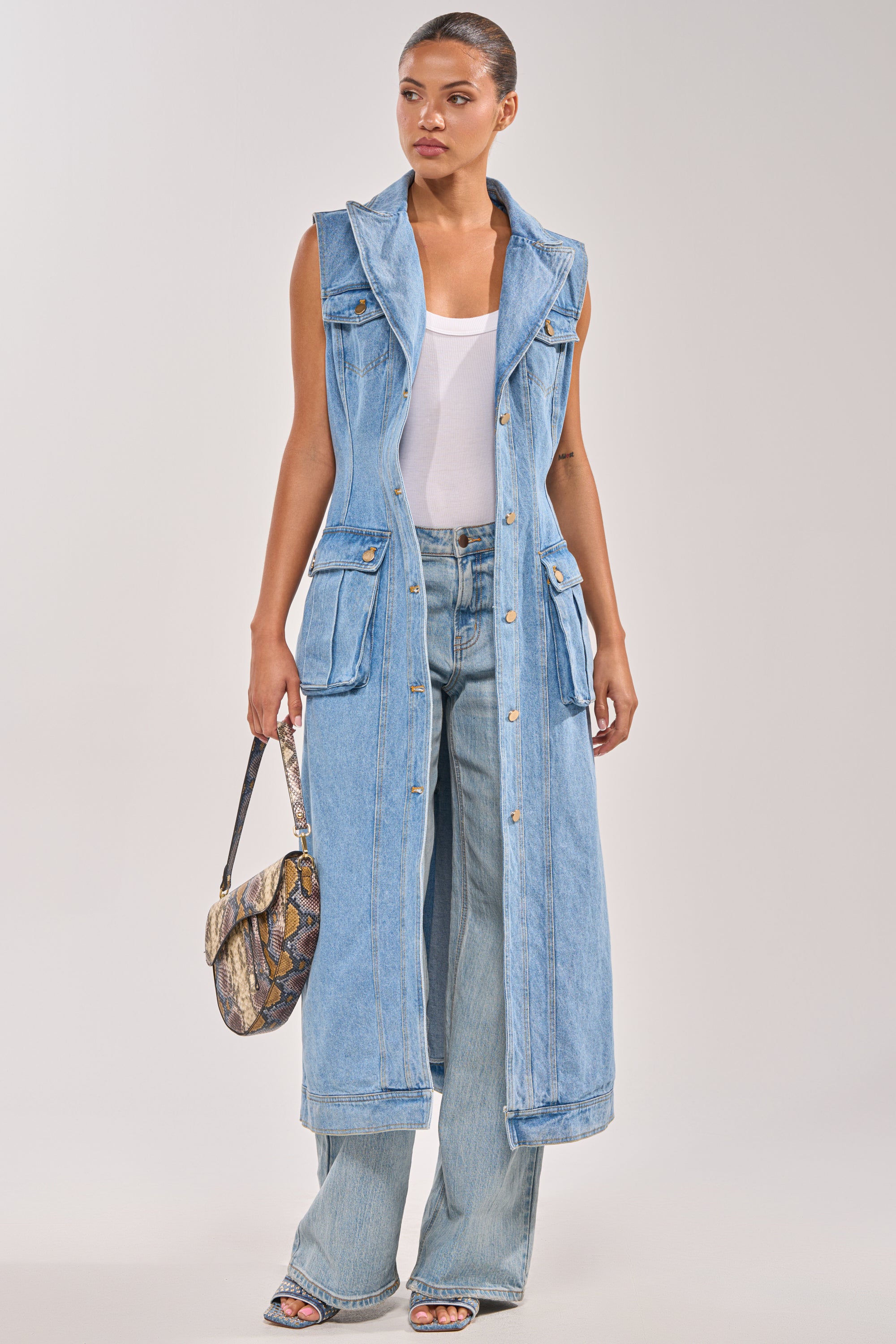 A woman wears the GOTTA DIP LONG DENIM VEST over a white tank top and wide-leg jeans, accessorizing with a small snakeskin-patterned handbag and open-toe heels, standing against a plain background. 1