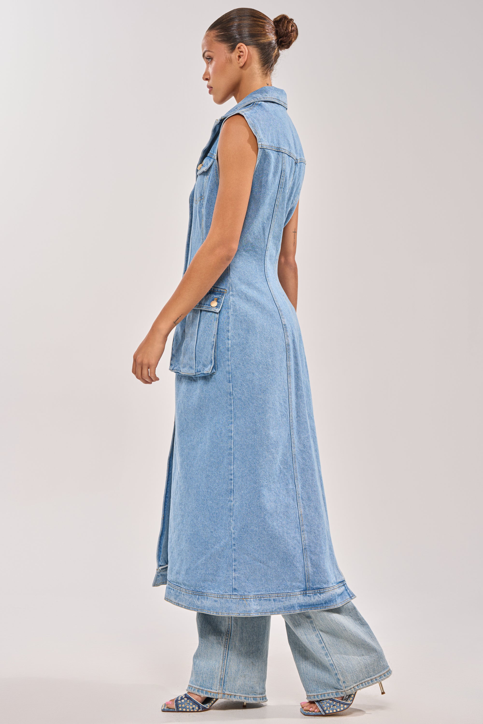 A woman with her hair in a bun stands in profile against a plain background, wearing the GOTTA DIP LONG DENIM VEST styled over matching wide-leg jeans and open-toe heeled sandals. 2
