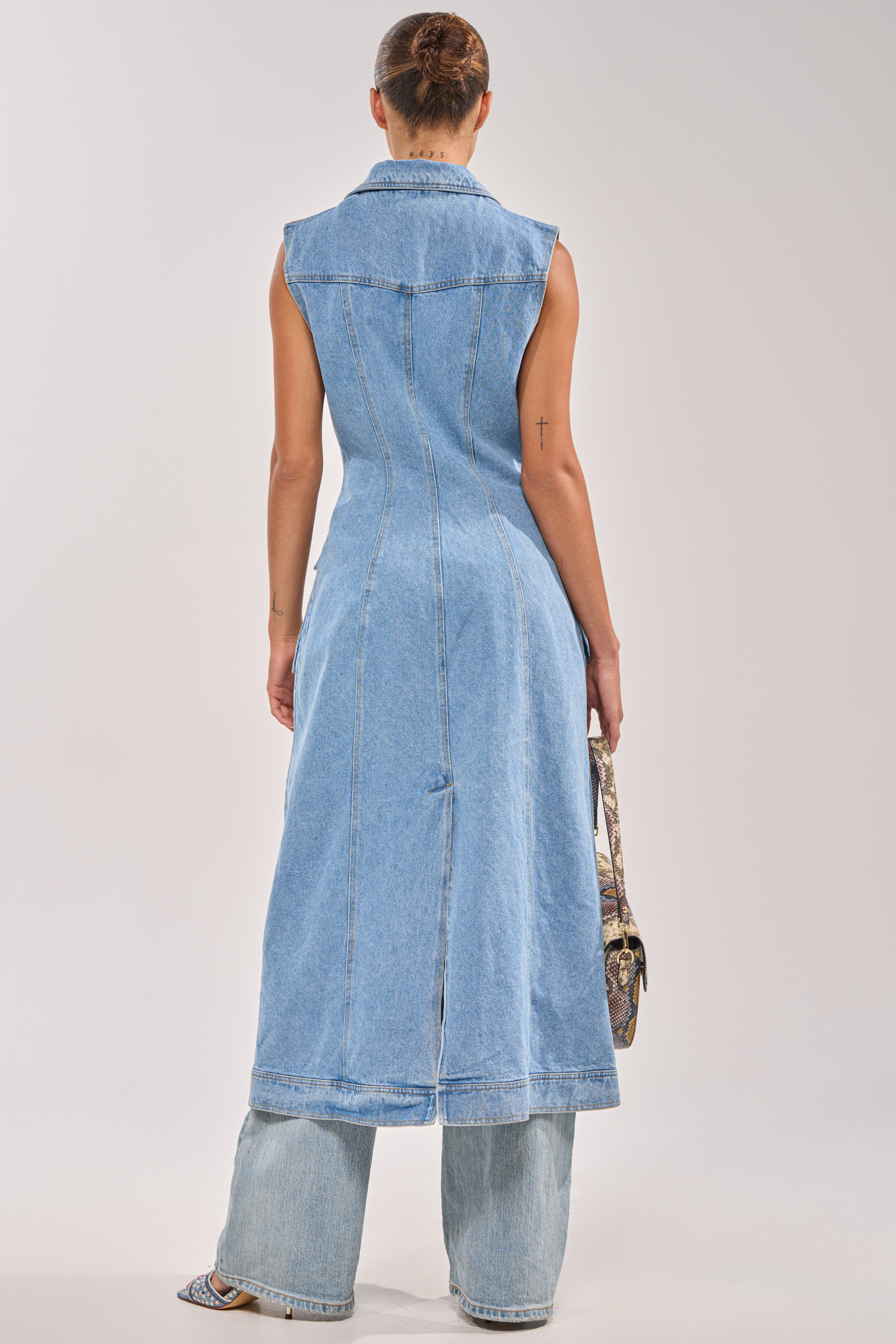 A person stands facing away, wearing the GOTTA DIP LONG DENIM VEST over wide-leg jeans. Their hair is in a neat bun, and they hold a patterned bag in their left hand. The background is plain and light-colored. 3
