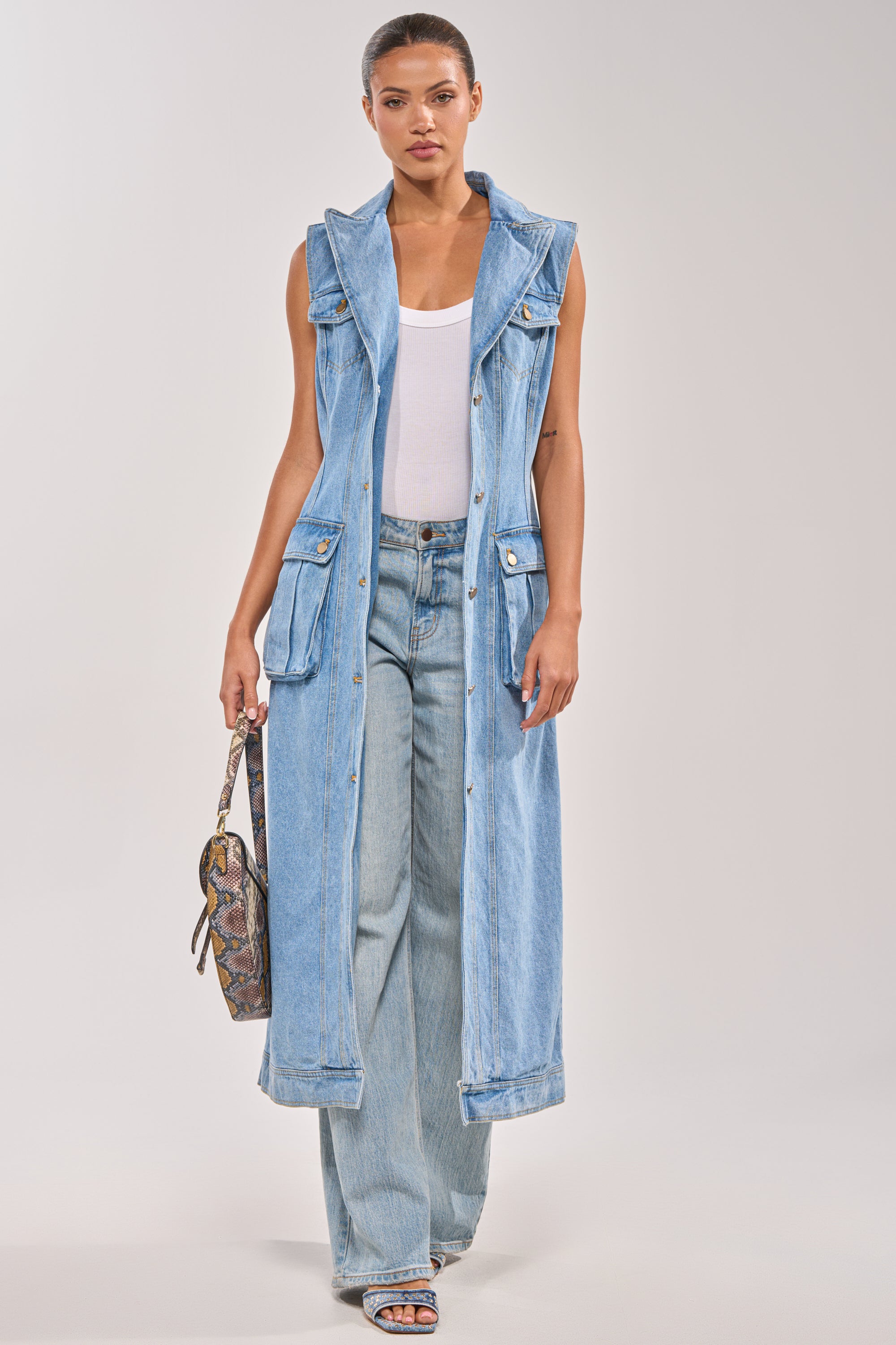 A woman models the GOTTA DIP LONG DENIM VEST over a white tank top and light blue wide-leg jeans, paired with blue sandals and a small patterned handbag, standing against a plain light background. 4