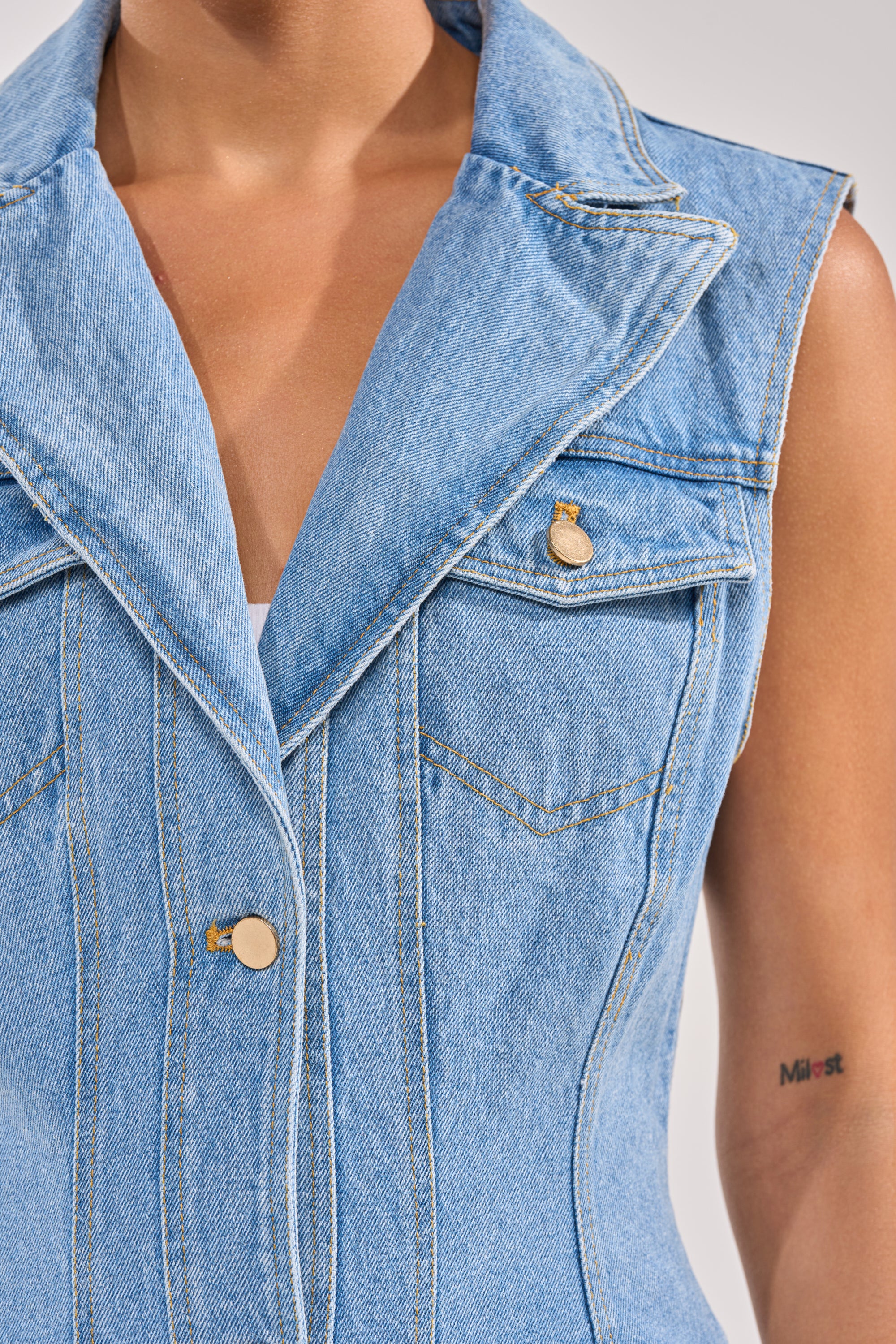 Close-up of a person wearing the GOTTA DIP LONG DENIM VEST, a sleeveless blue denim cargo vest with gold buttons. A small colorful tattoo appears on their upper arm near the word "Mist." The background is plain and neutral. 5