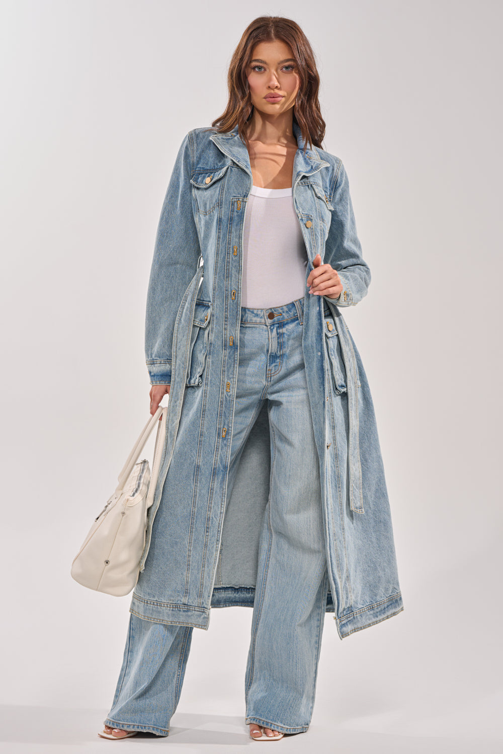 A woman poses confidently in the CAMILLA DENIM TRENCH over a white top and wide-leg jeans, holding a white handbag against a plain, light background. 4