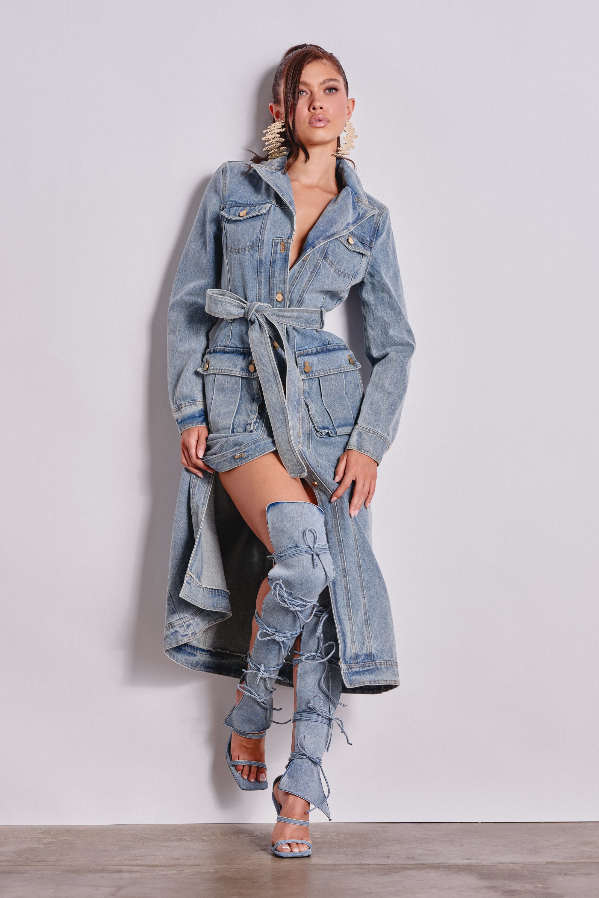 A woman poses confidently against a white wall wearing the CAMILLA DENIM TRENCH with a belt, paired with matching high lace-up denim boots and bold statement earrings, one knee slightly bent. 0