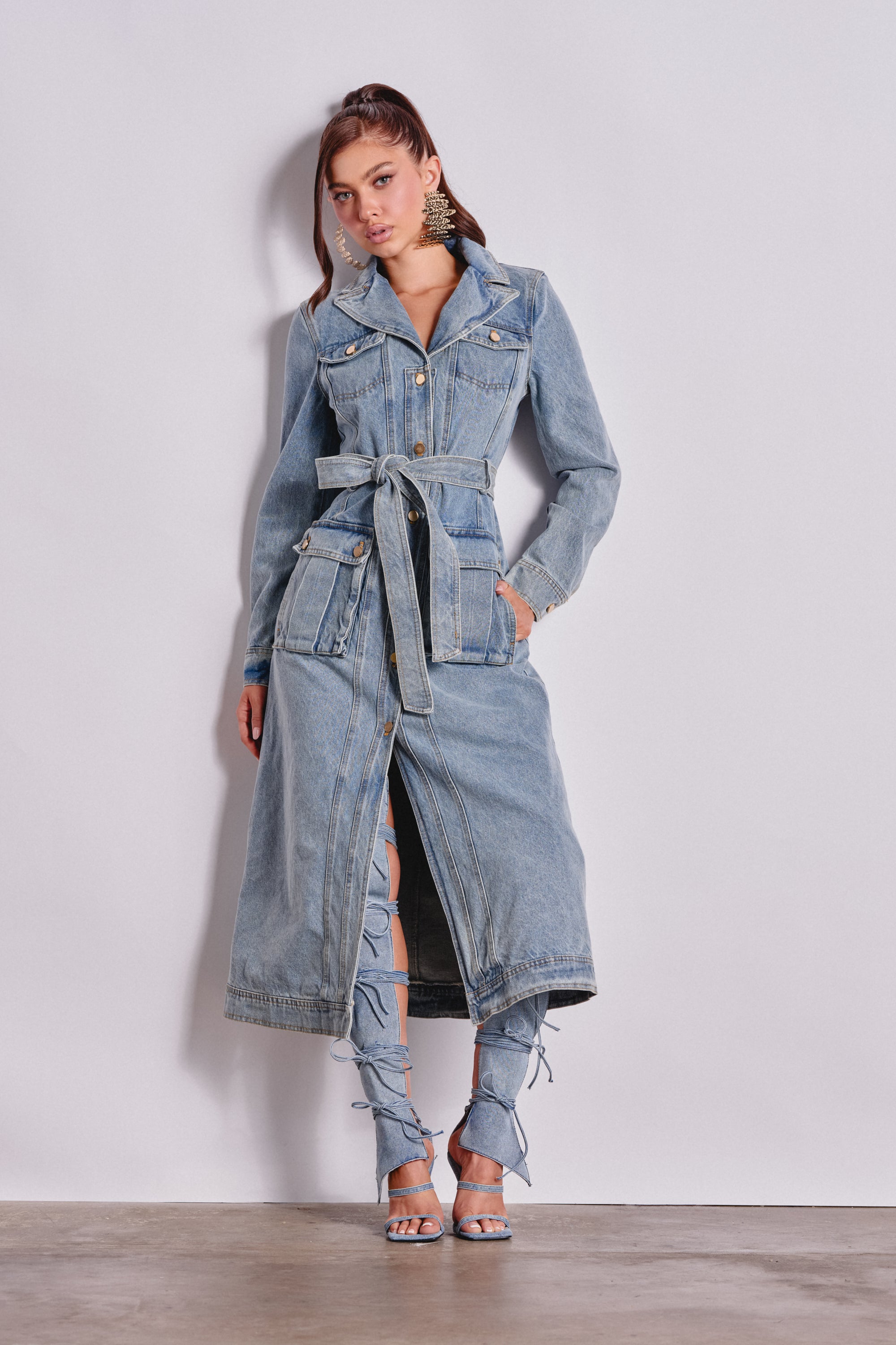 A woman poses against a white wall wearing the CAMILLA DENIM TRENCH over matching slit denim pants and strappy heeled sandals. She accessorizes with statement earrings and has her hair partially tied back. 1