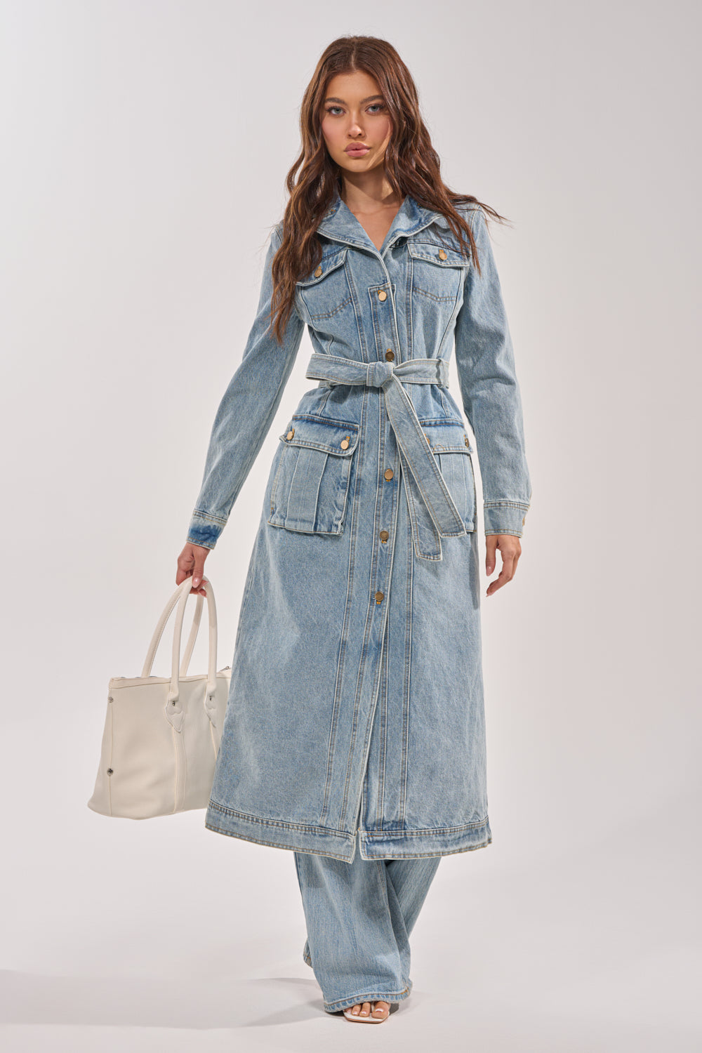 A woman wears the CAMILLA DENIM TRENCH with cargo pockets over matching jeans and open-toed shoes, carrying a large white handbag against a plain light backdrop. 6