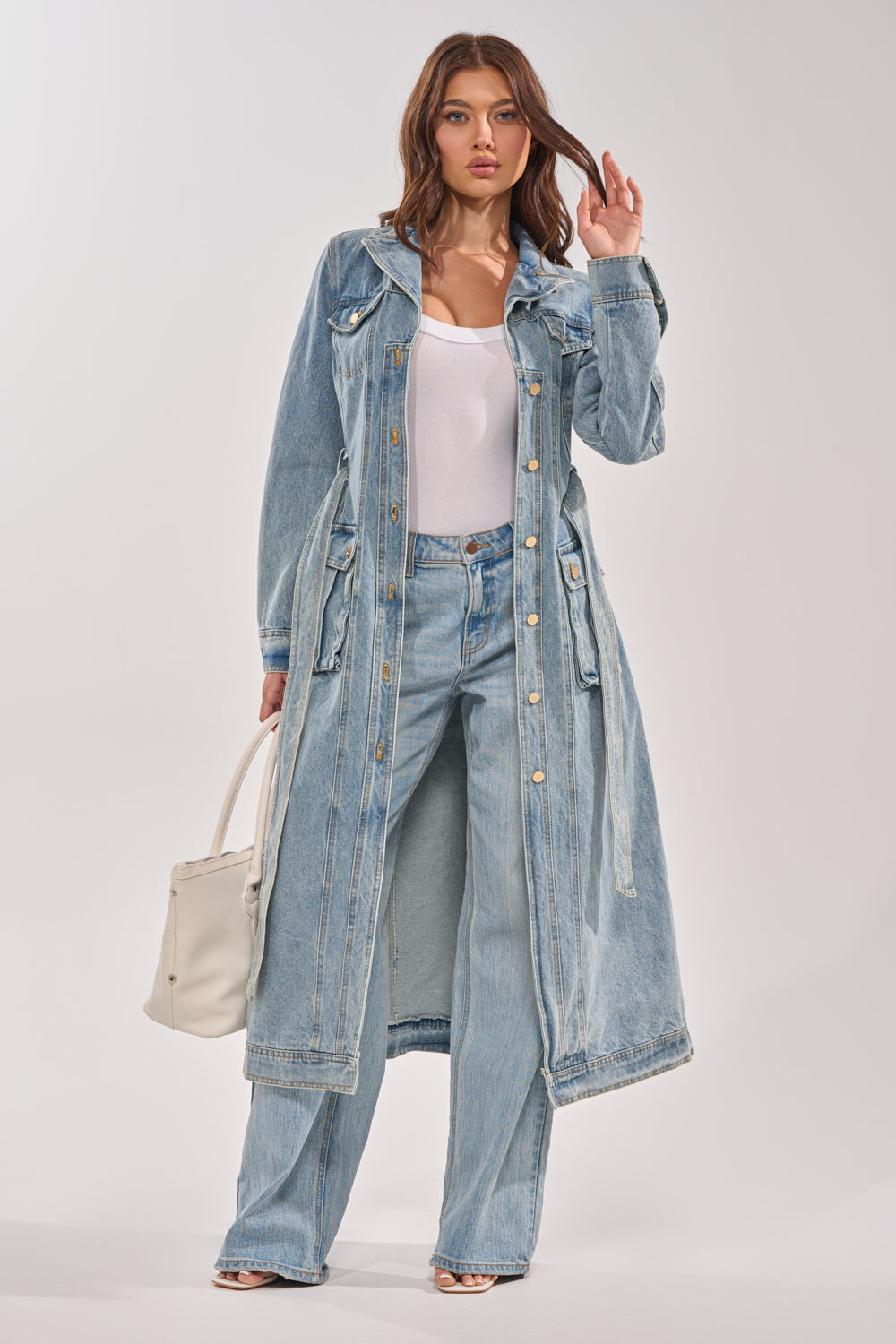 A woman wearing the CAMILLA DENIM TRENCH over a white top and light blue jeans, holding a white handbag, stands against a plain light background. 7