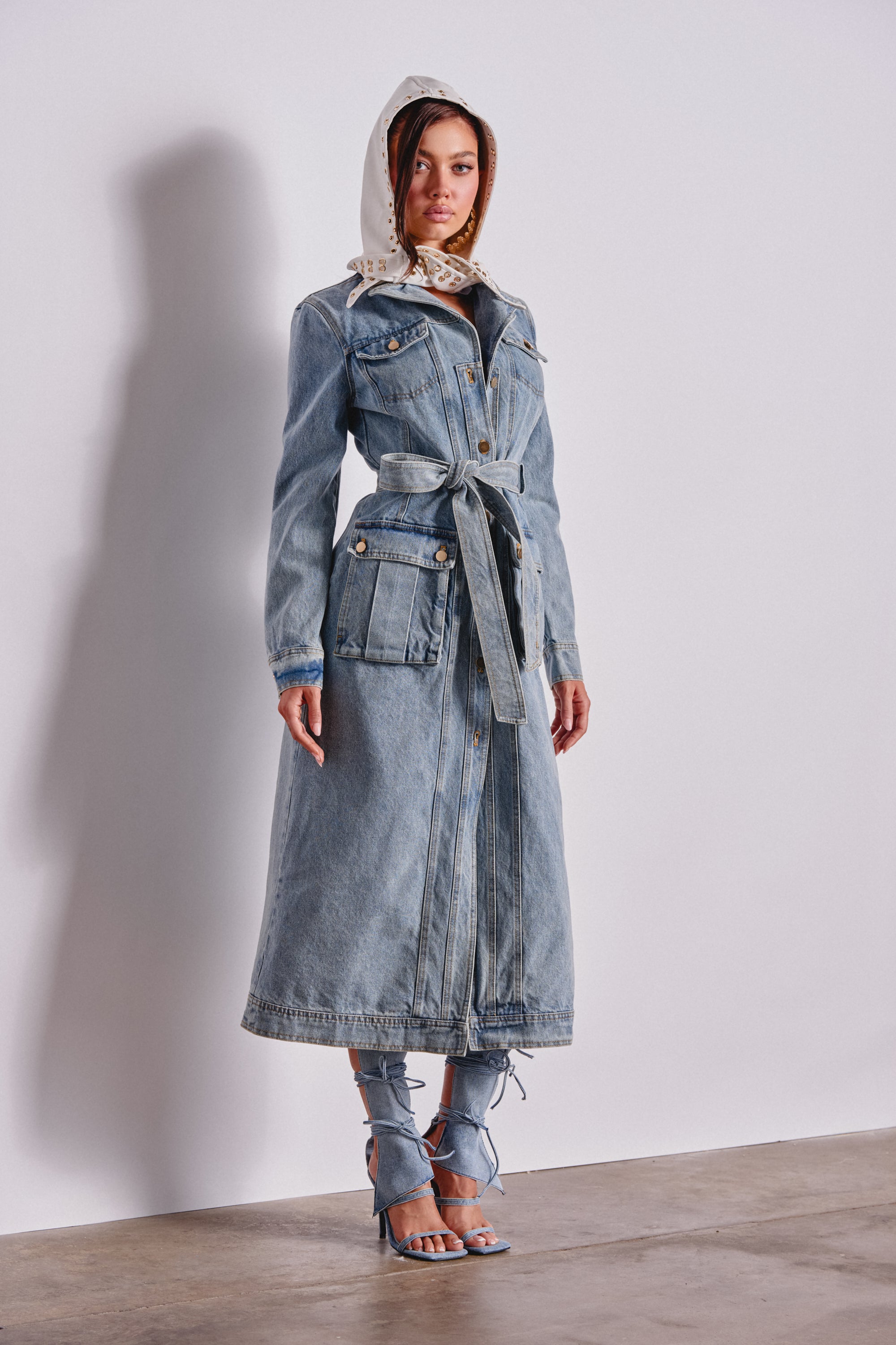 A woman poses against a white wall in the CAMILLA DENIM TRENCH, styled with denim-wrapped heeled sandals and a white bandana hood; she stands with one hand by her side and the other slightly forward. 3