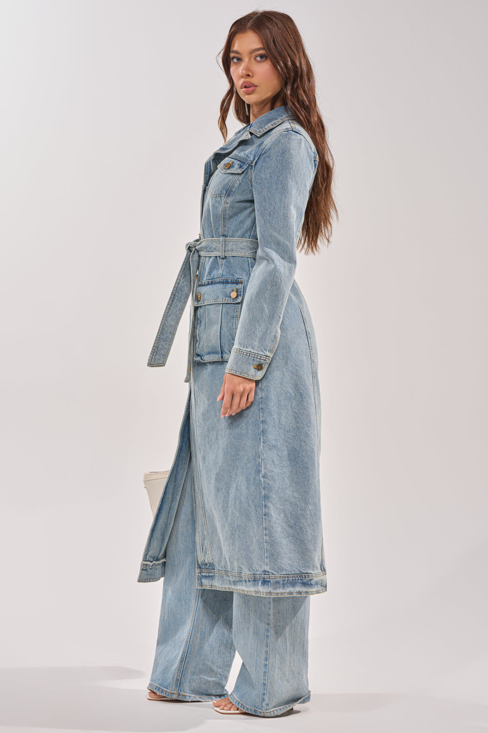 A woman with long brown hair models the CAMILLA DENIM TRENCH—a light blue belted denim coat—paired with matching wide-leg jeans and open-toe heels, standing sideways against a plain light backdrop. 8