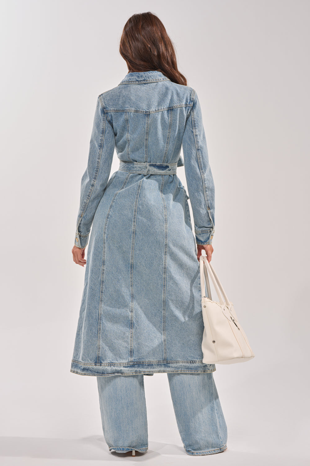 A woman with long brown hair stands facing away, wearing the CAMILLA DENIM TRENCH over wide-leg jeans. She carries a white handbag, set against a plain light background. 9