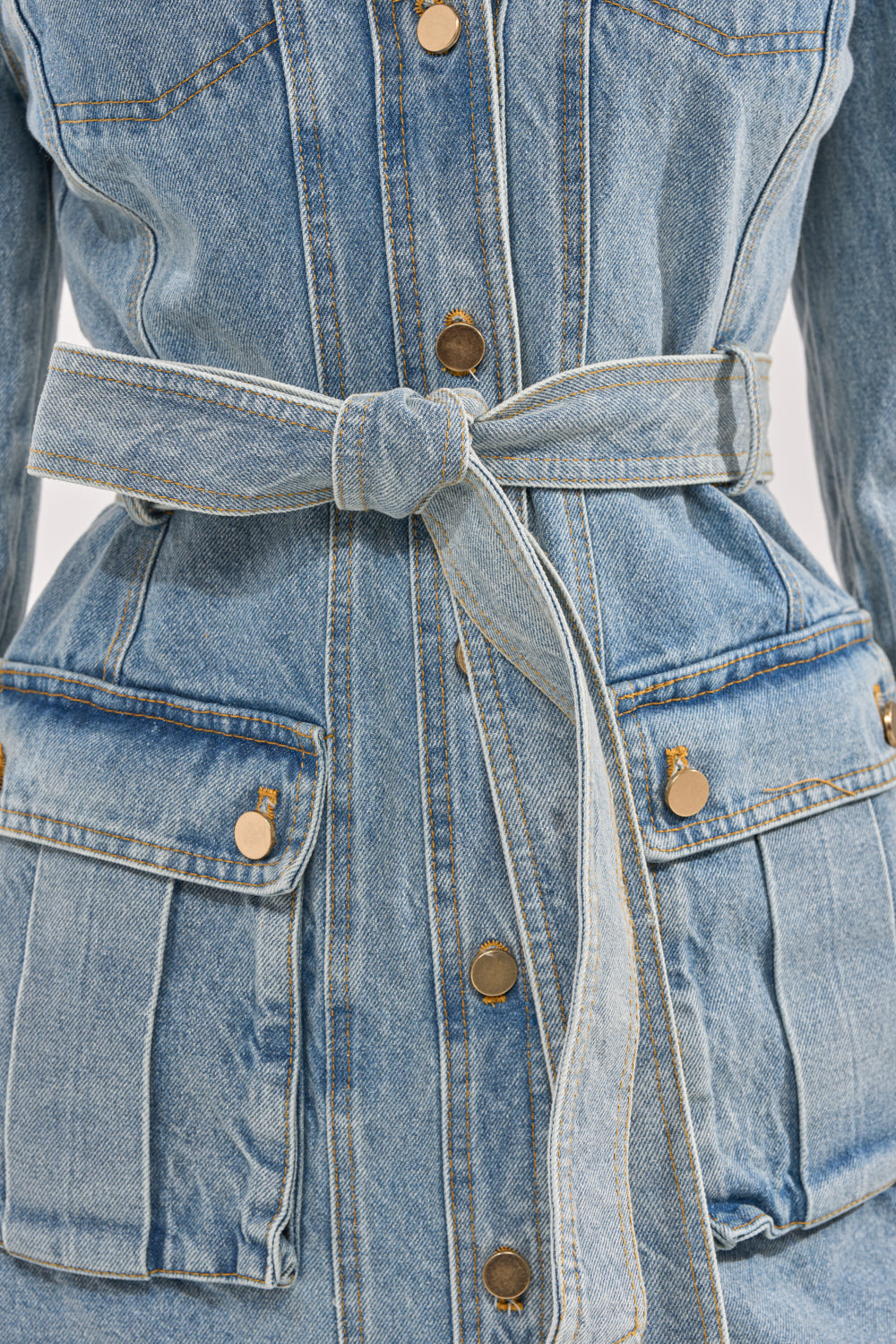Close-up of a person wearing the CAMILLA DENIM TRENCH, featuring a belted waist, gold buttons, and two large front pockets for a modern, fitted utility-jacket look. 10