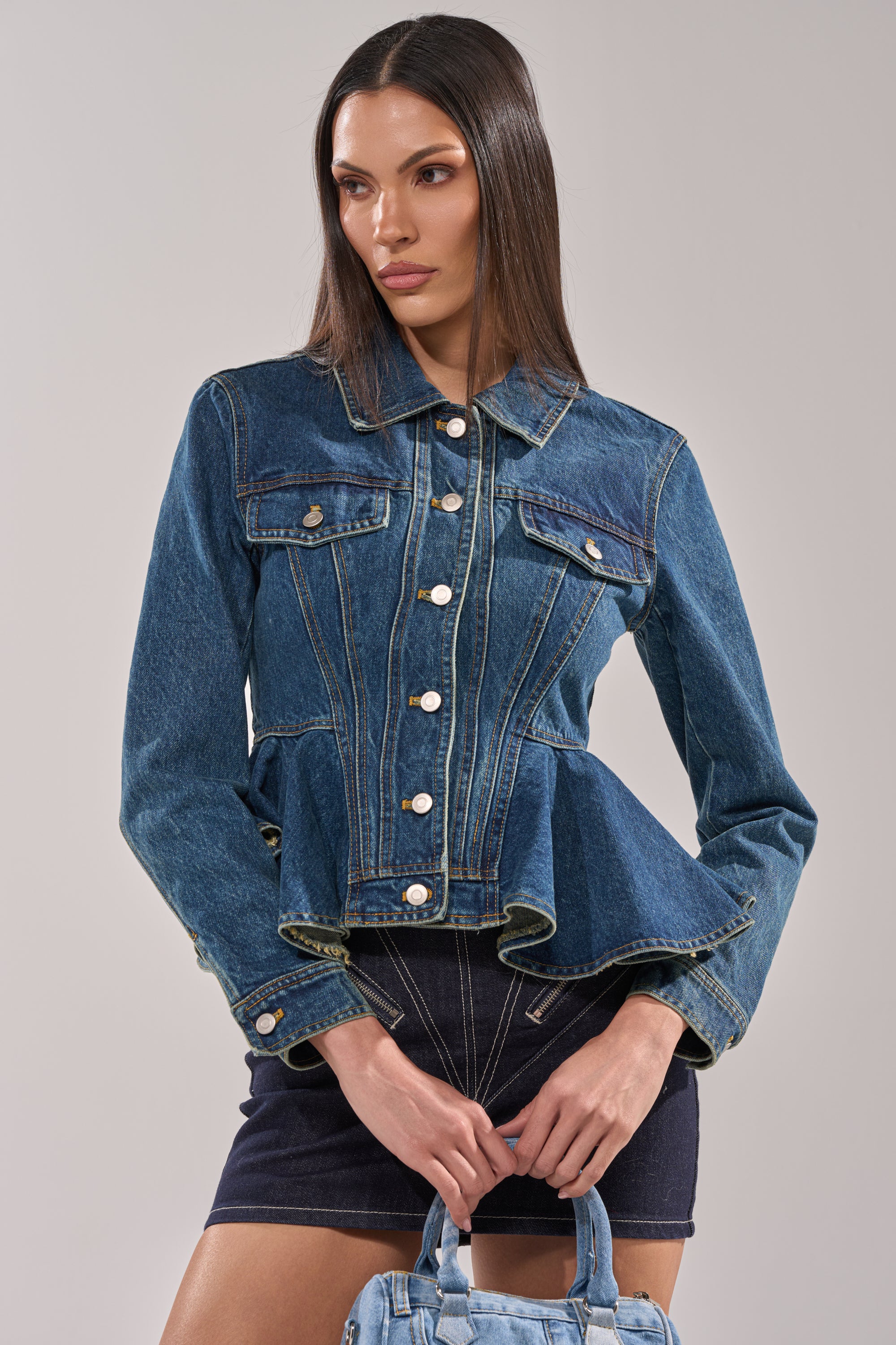 A woman wears the POP OUT PEPLUM DENIM JACKET with a peplum hem and silver buttons, styled with a dark denim skirt and small denim handbag, standing against a plain light gray background. 0