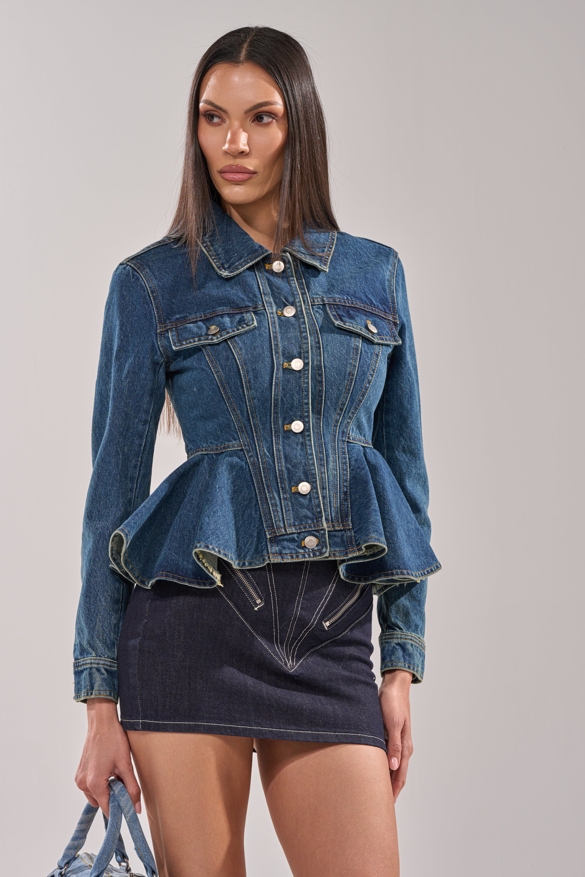 A woman models the POP OUT PEPLUM DENIM JACKET with silver buttons, paired with a high-waisted denim mini skirt and matching bag against a plain background—a chic addition to any women's jacket collection. 2