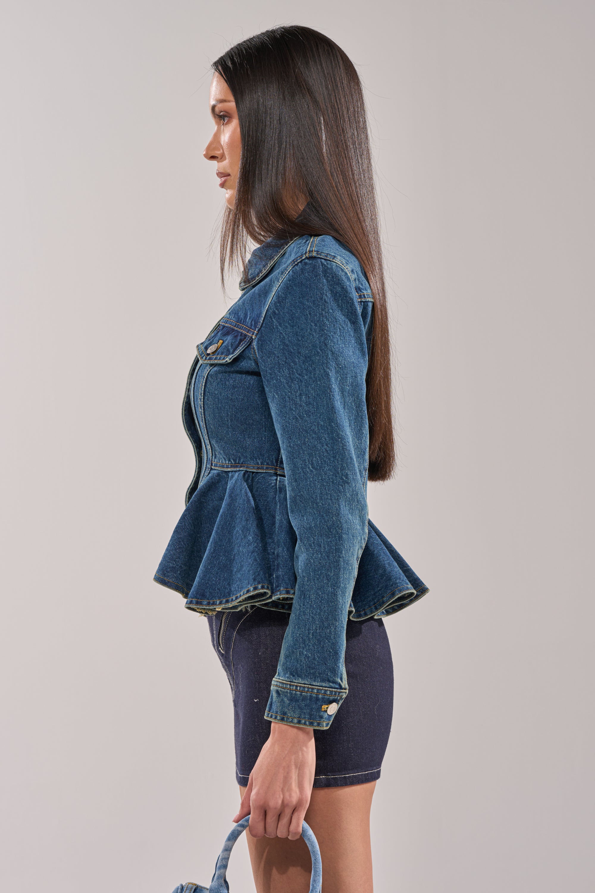 A woman with straight, long brown hair stands in profile wearing the POP OUT PEPLUM DENIM JACKET and a dark skirt, holding a blue handbag against a plain light backdrop—an ideal casual layering piece. 3