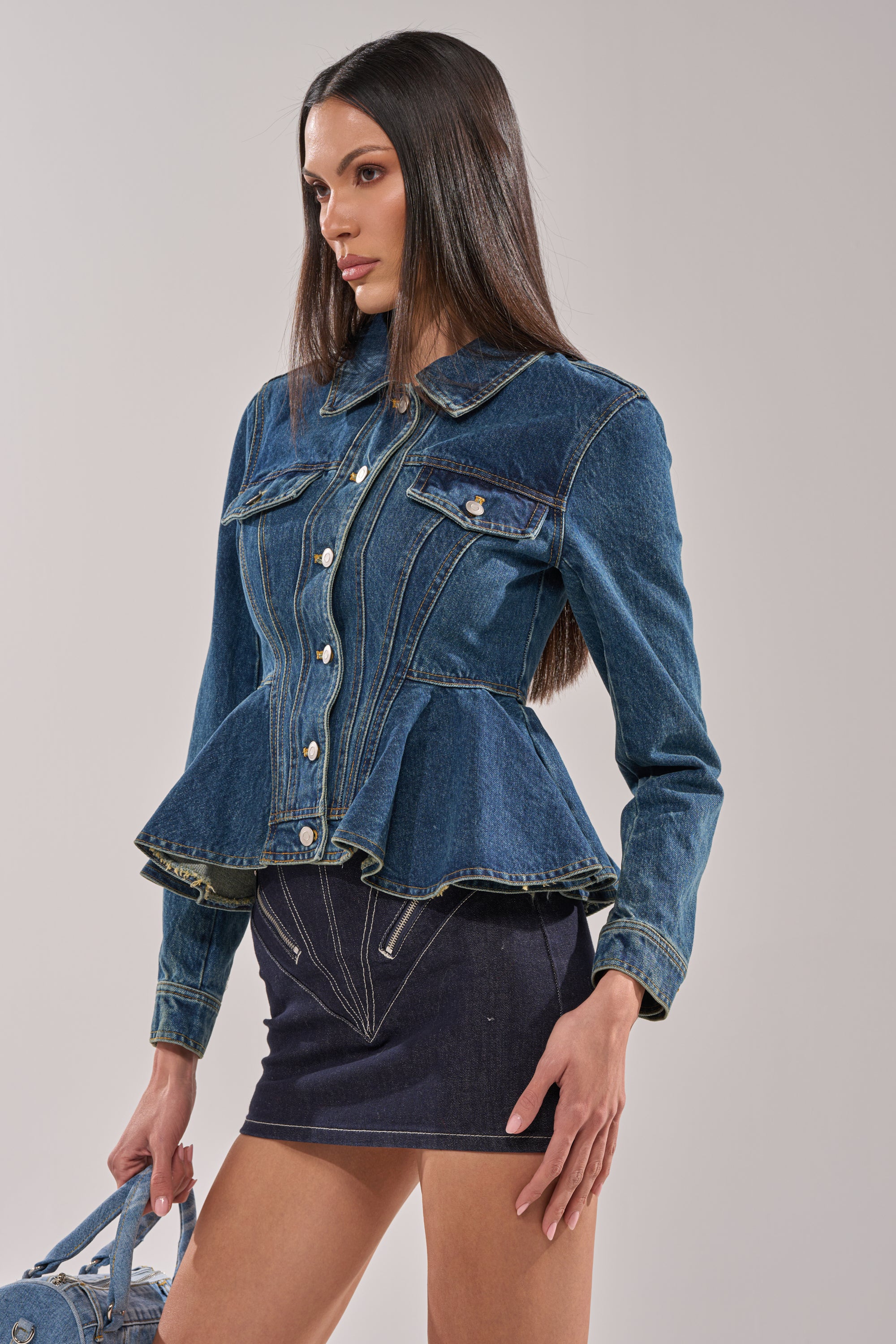 A woman wears the POP OUT PEPLUM DENIM JACKET with a button-up front and dark denim mini skirt, holding a light blue handbag against a plain light background. 4