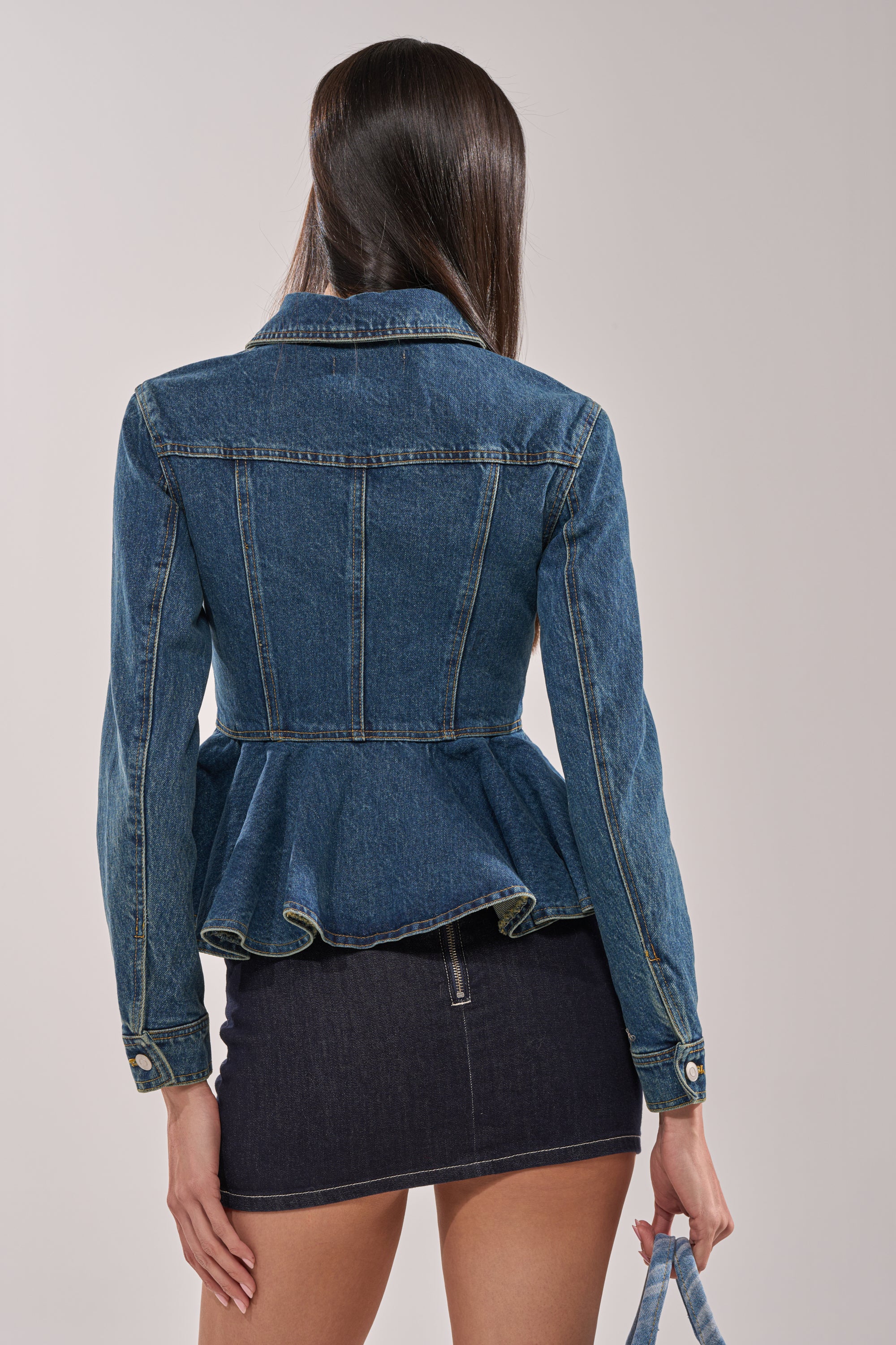 A woman with long straight hair faces away, showcasing the POP OUT PEPLUM DENIM JACKET styled with a dark denim mini skirt and matching denim bag, creating an effortlessly chic layered look. 5