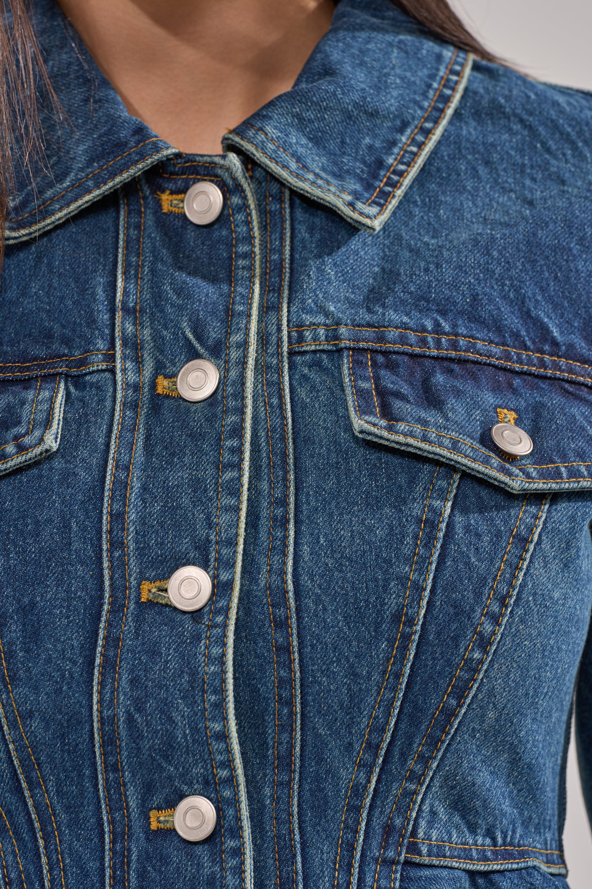Close-up of a person's upper body wearing the POP OUT PEPLUM DENIM JACKET, featuring metal buttons, yellow stitching, chest pockets, and detailed seams near the collar. 8