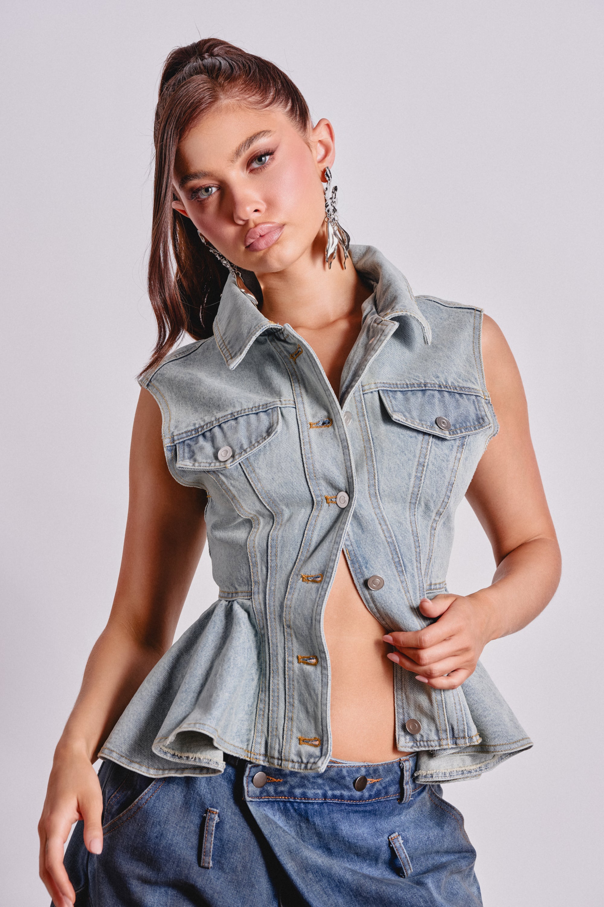 A woman models the FORGET ABOUT YOU PEPLUM DENIM VEST, a sleeveless light-wash denim piece with a flared hem and high collar, styled with dark blue jeans. She wears her long brown hair in a ponytail and pairs it with bold statement earrings. 1
