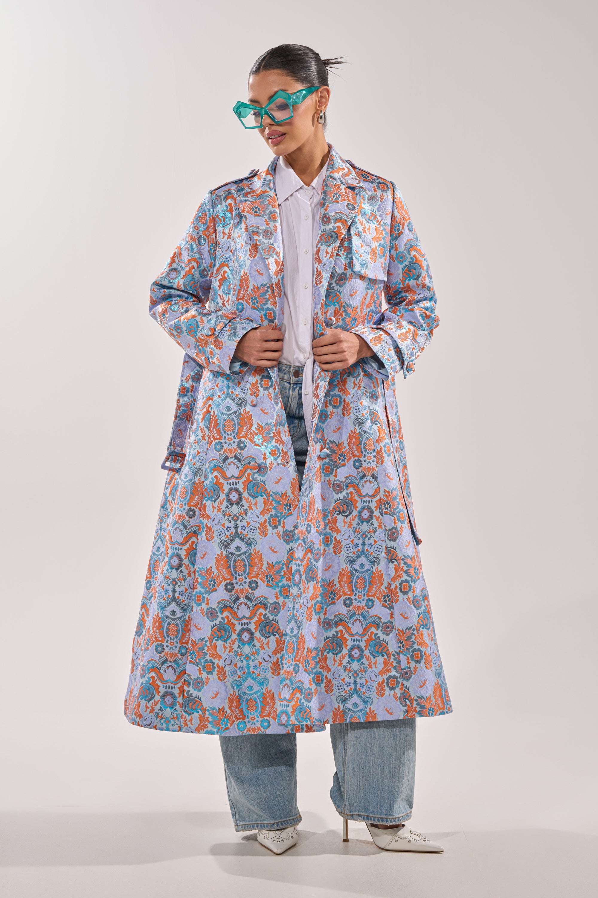 A person stands against a plain light background wearing the ST. GERMAIN BROCADE TRENCH, turquoise glasses, a white shirt, blue jeans, and white heels. 0