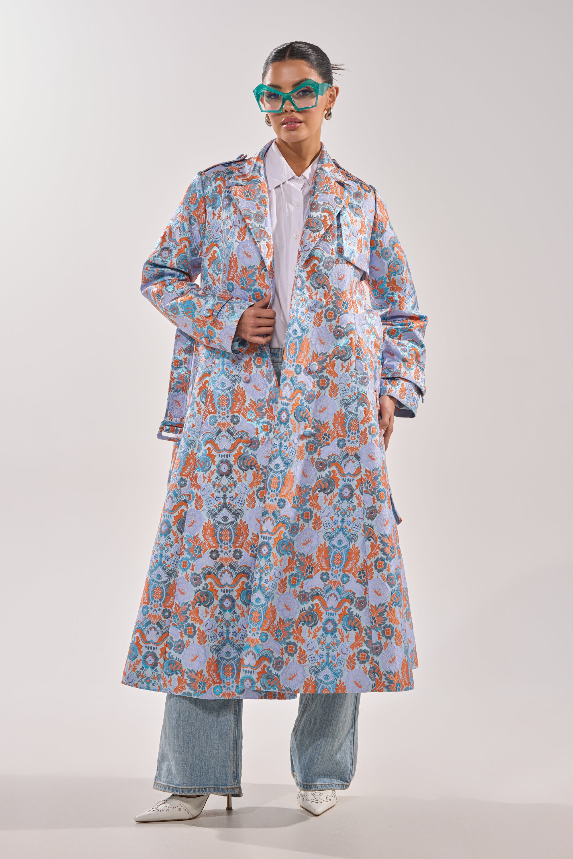 A woman wearing teal glasses styles the ST. GERMAIN BROCADE TRENCH over a white shirt and light blue jeans with white pointed-toe heels, standing confidently against a plain background. 2
