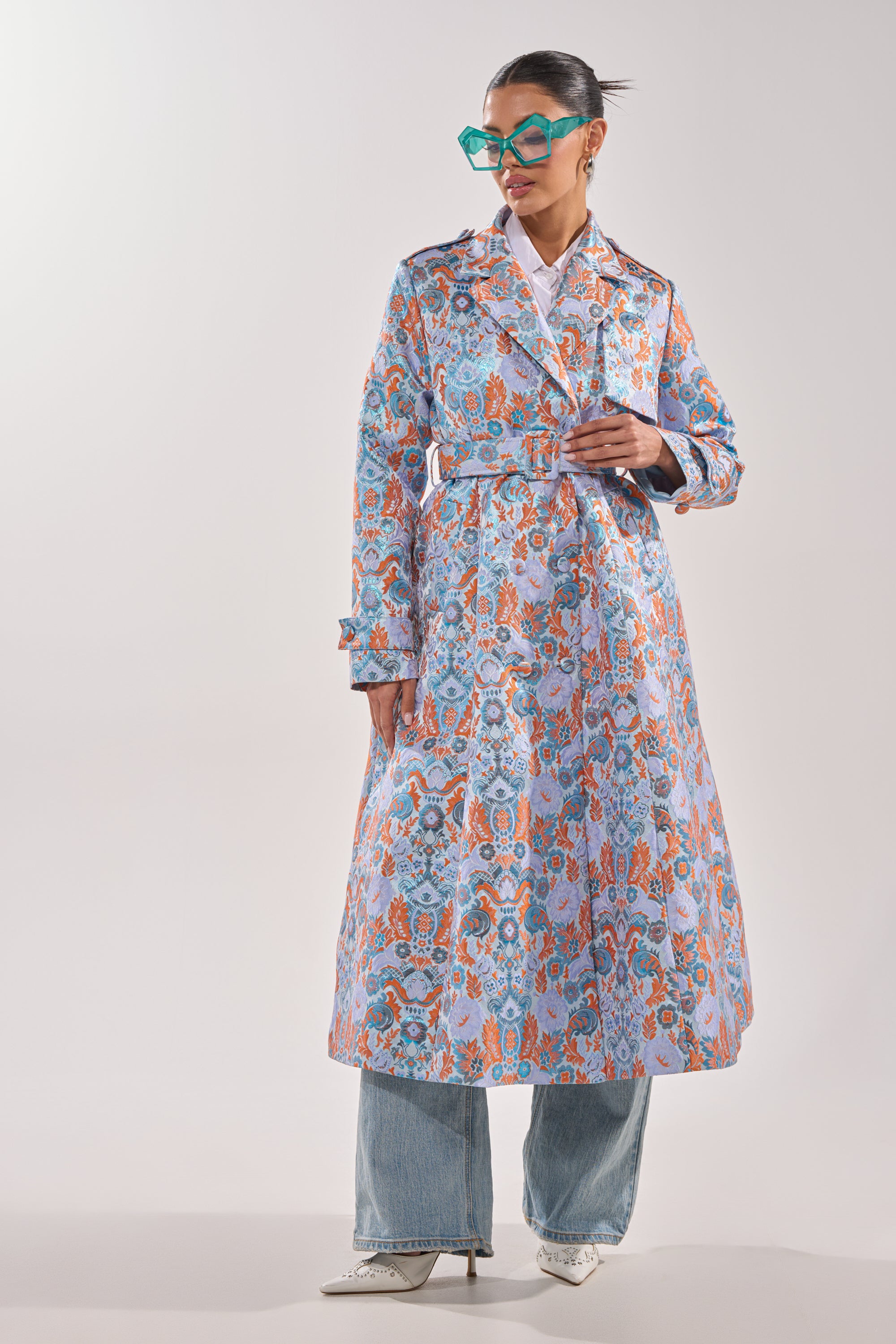 A woman poses confidently against a plain light background wearing the ST. GERMAIN BROCADE TRENCH, featuring pastel blue, orange, and pink designs, styled with wide-leg jeans, white boots, and oversized teal glasses. 3