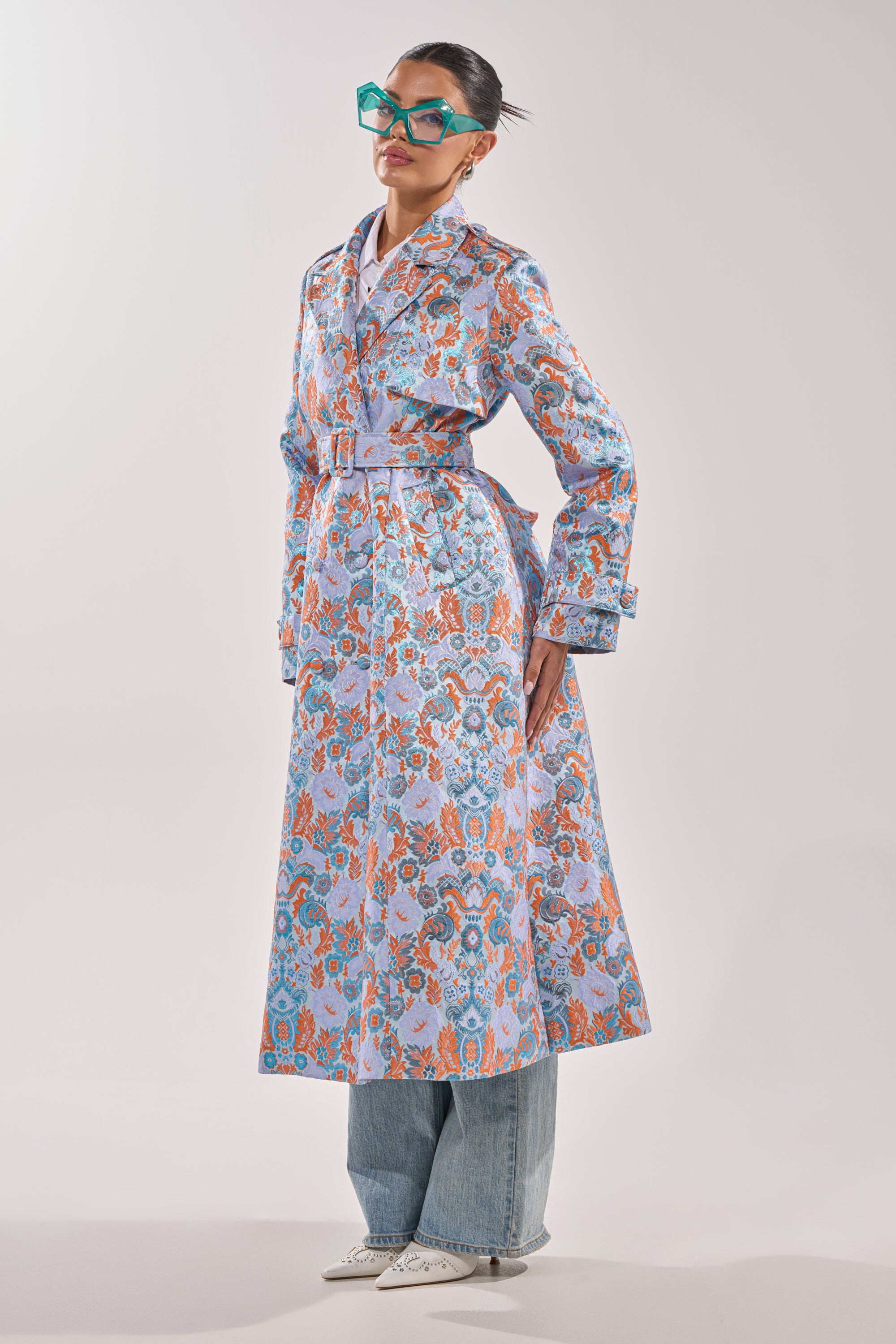 A woman wearing the ST. GERMAIN BROCADE TRENCH over blue jeans, with pointed white shoes and oversized turquoise sunglasses, stands against a plain light background. 4