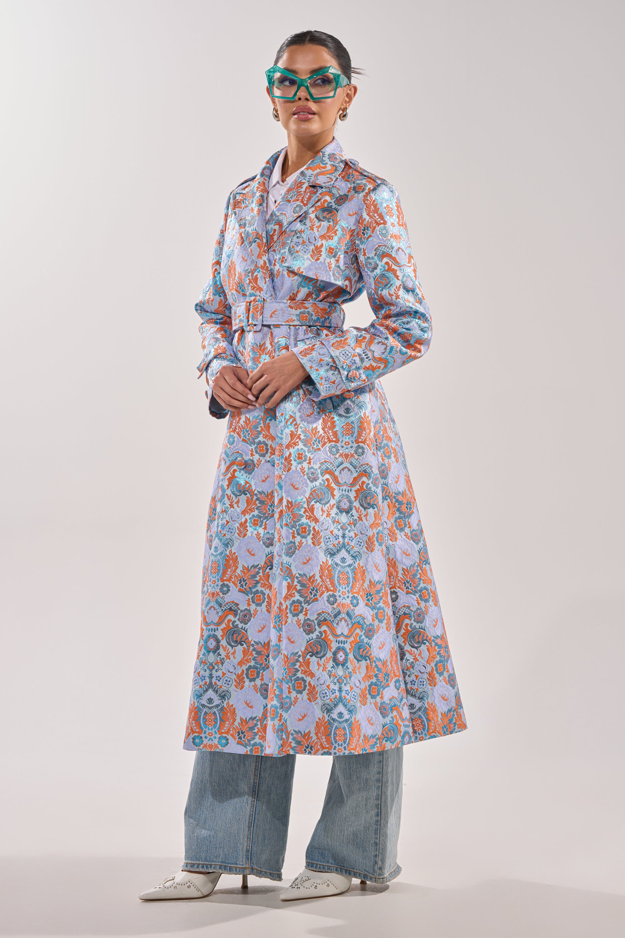 Wearing the ST. GERMAIN BROCADE TRENCH over blue jeans, cat-eye sunglasses, and white pointed shoes, a person stands against a plain light background. 5