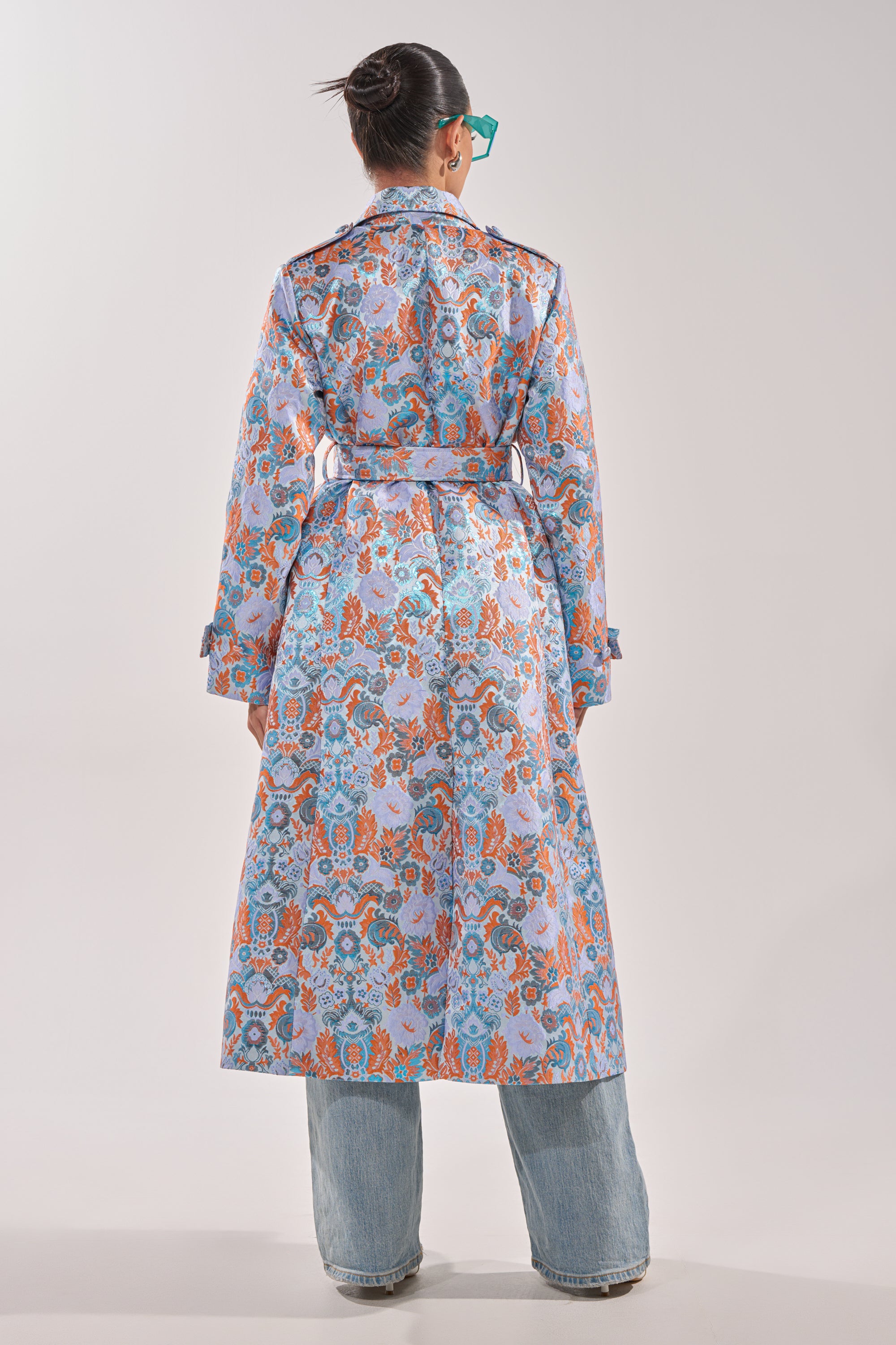 A person with a bun and teal glasses models the ST. GERMAIN BROCADE TRENCH, its vibrant floral pattern standing out over wide-legged blue jeans against a simple, light background. 6