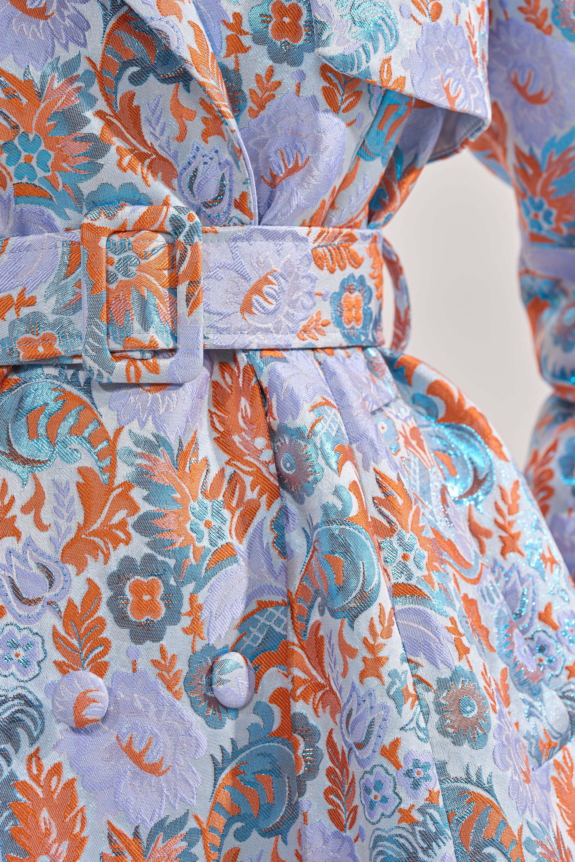 Close-up of the ST. GERMAIN BROCADE TRENCH, featuring intricate orange, turquoise, and lavender floral patterns on textured fabric, detailed with buttons and a matching belt with a rectangular buckle. 7
