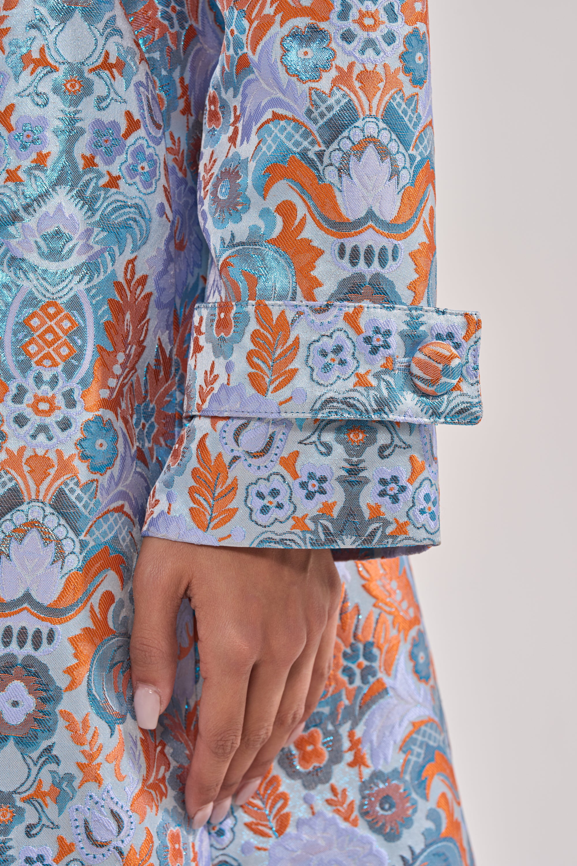 A close-up of a hand resting by the side, wearing the ST. GERMAIN BROCADE TRENCH in blue, orange, and silver with intricate floral and geometric patterns and wide cuff detail. 8