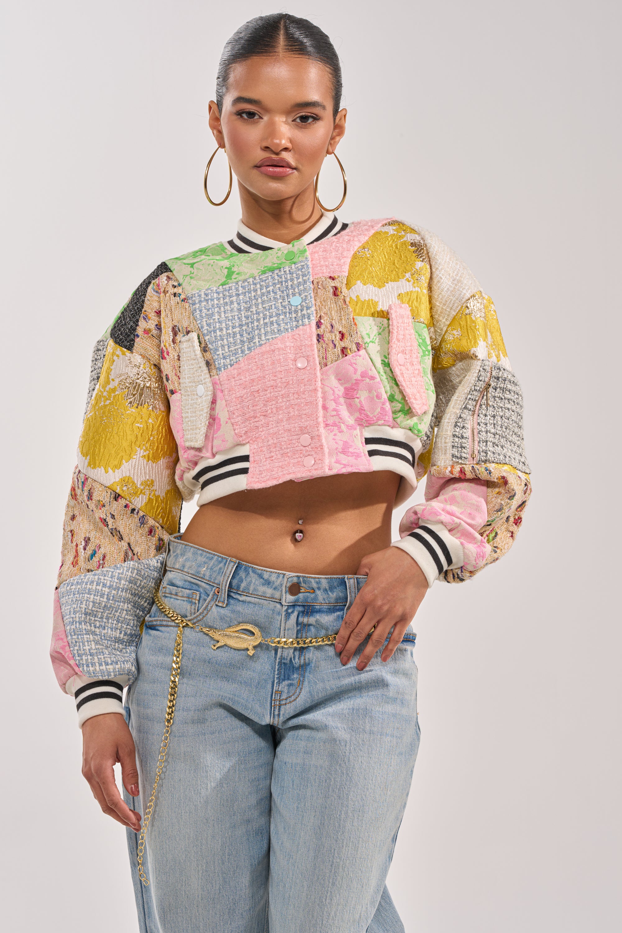 A woman stands confidently against a plain background in a statement streetwear look, wearing the ESTELLE PATCHWORK BOMBER with striped cuffs and light blue jeans featuring a gold chain belt. 0
