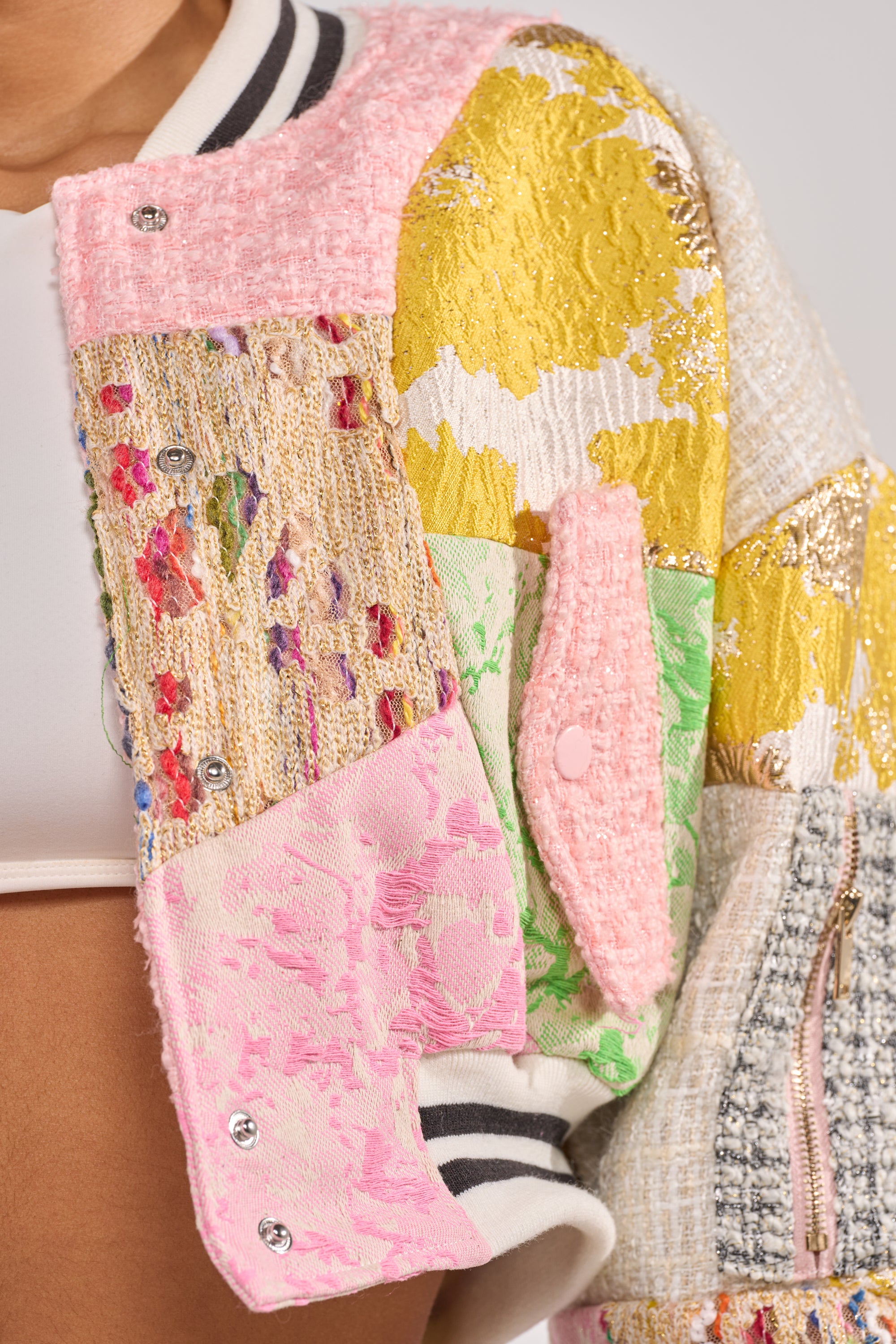 A close-up of the ESTELLE PATCHWORK BOMBER, a statement streetwear jacket with textured fabrics in pink, gold, green, and cream over a white crop top. Features include a ribbed collar and snap buttons. 9
